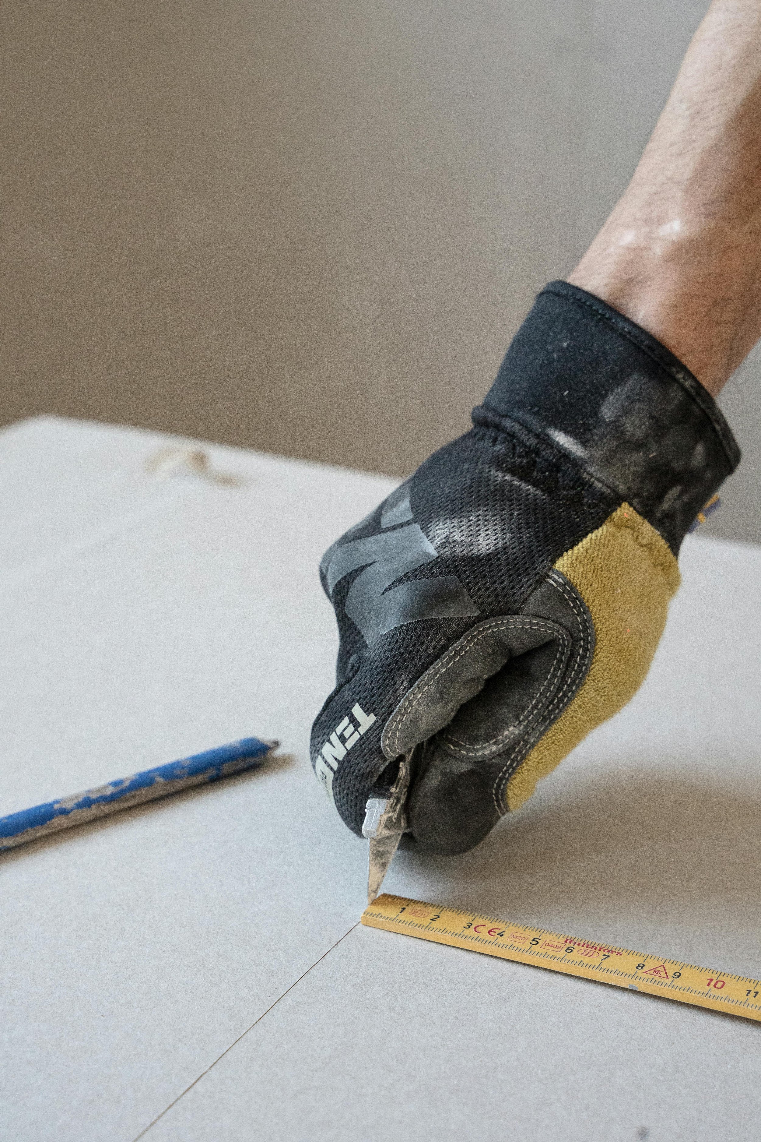 A close-up of a gloved hand holding a measuring tape against a plain surface, with a flat object and a pen nearby on the table.