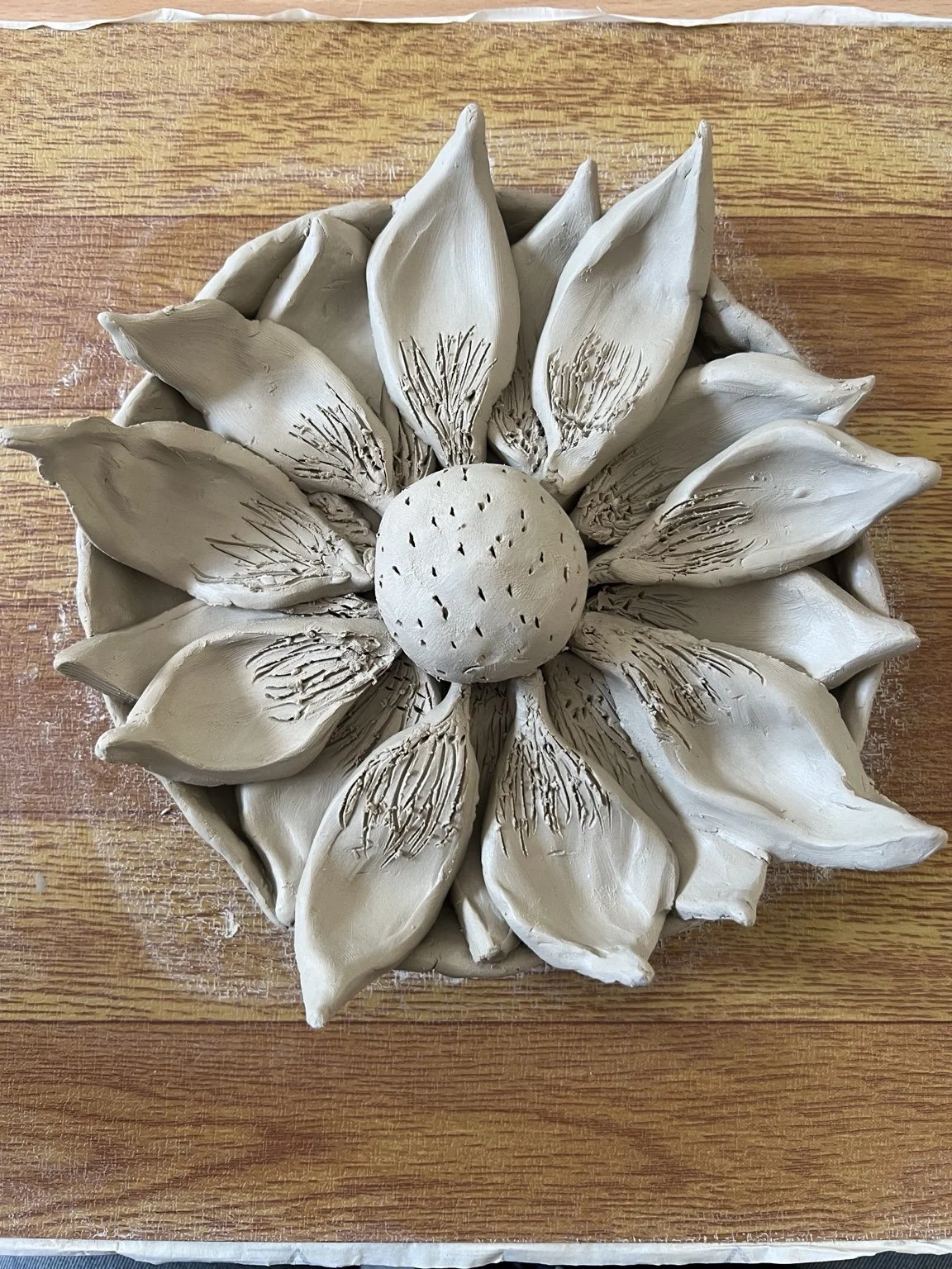 Unfinished ceramic sculpture of a sunflower with petals around a round seed head, placed on a wooden surface.