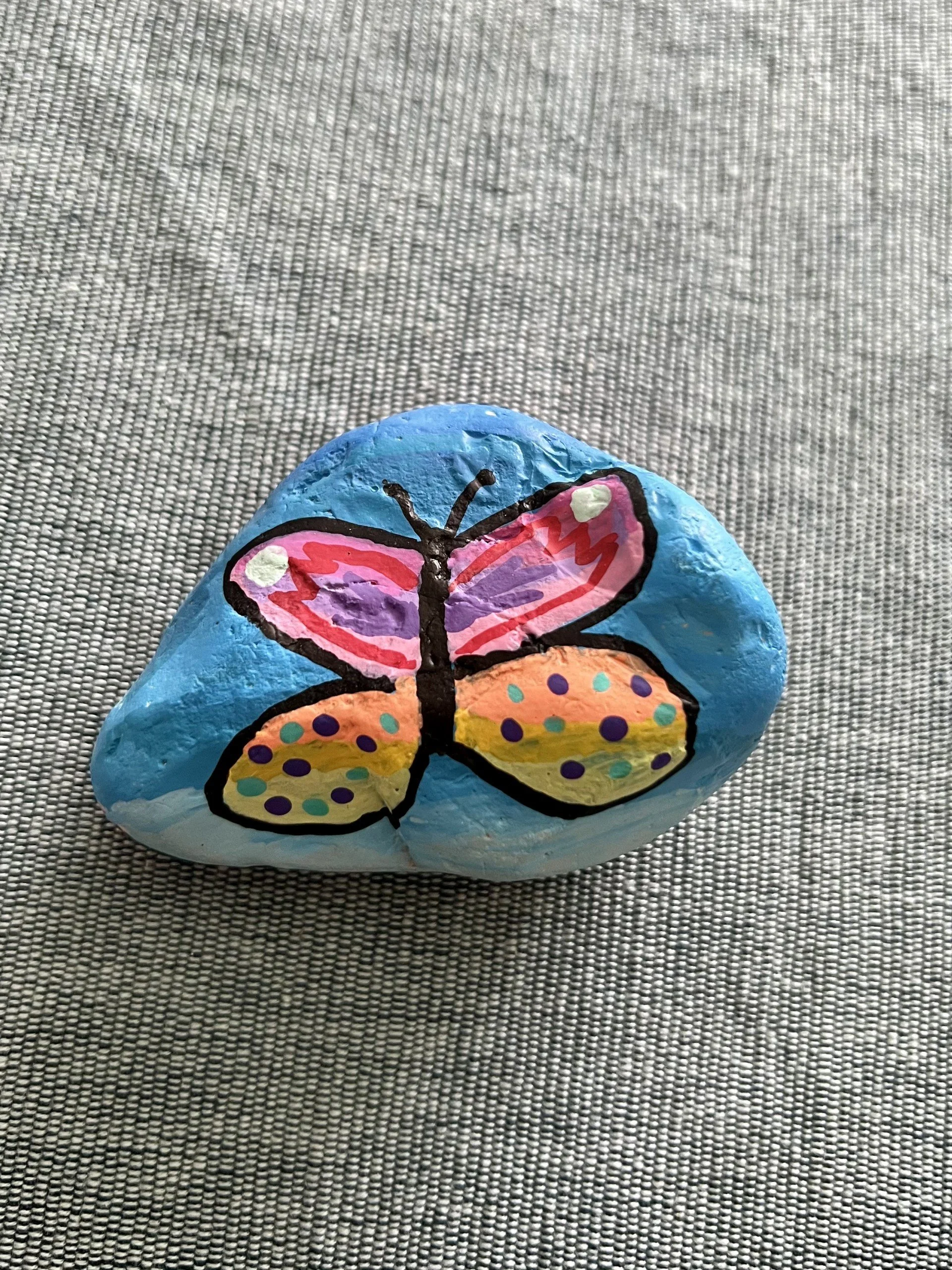 Painted rock with a colorful butterfly design, placed on a gray textured surface.