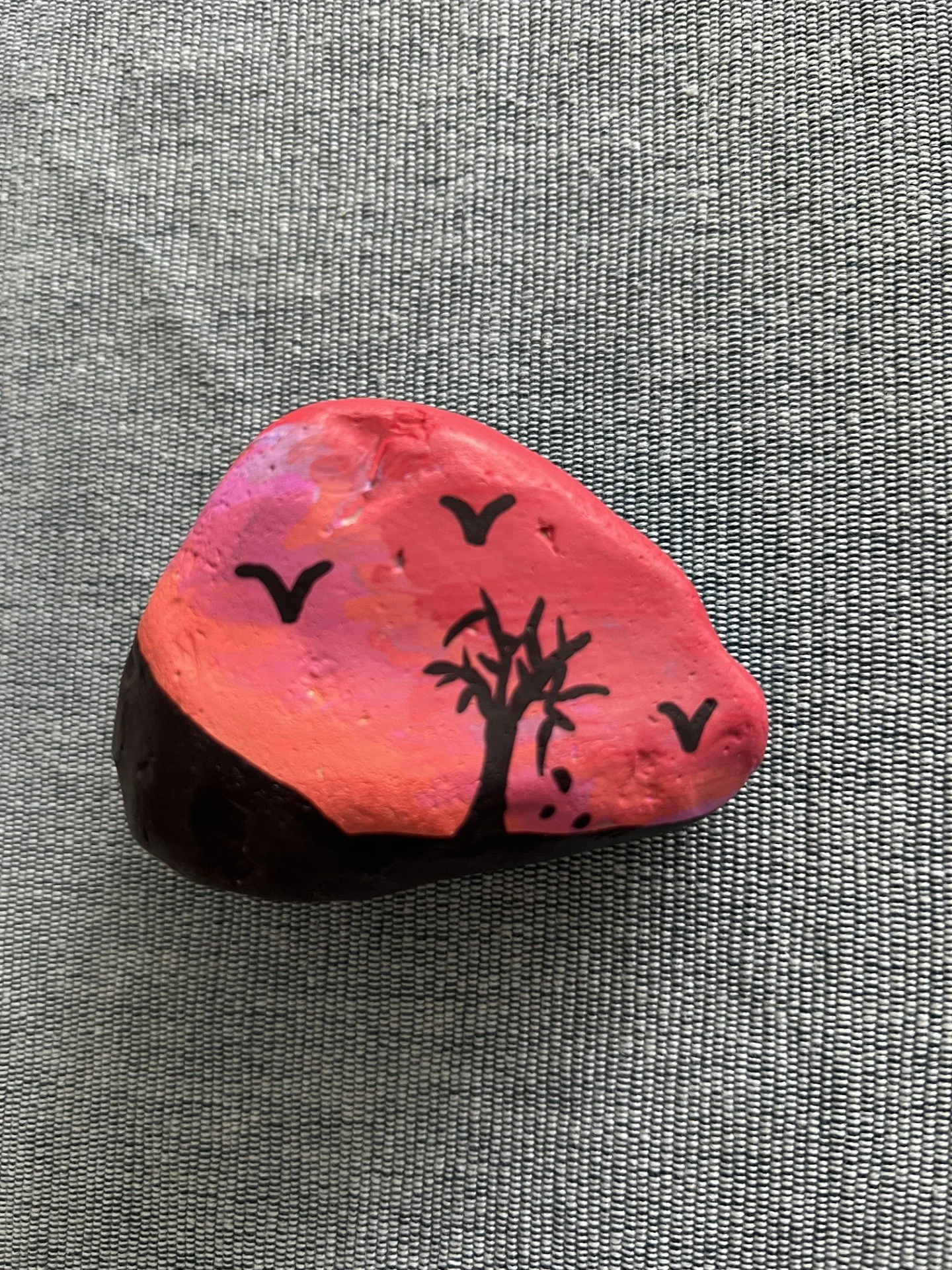 A painted rock with a pink and orange background featuring a black tree silhouette and flying bird shapes, placed on a textured gray surface.