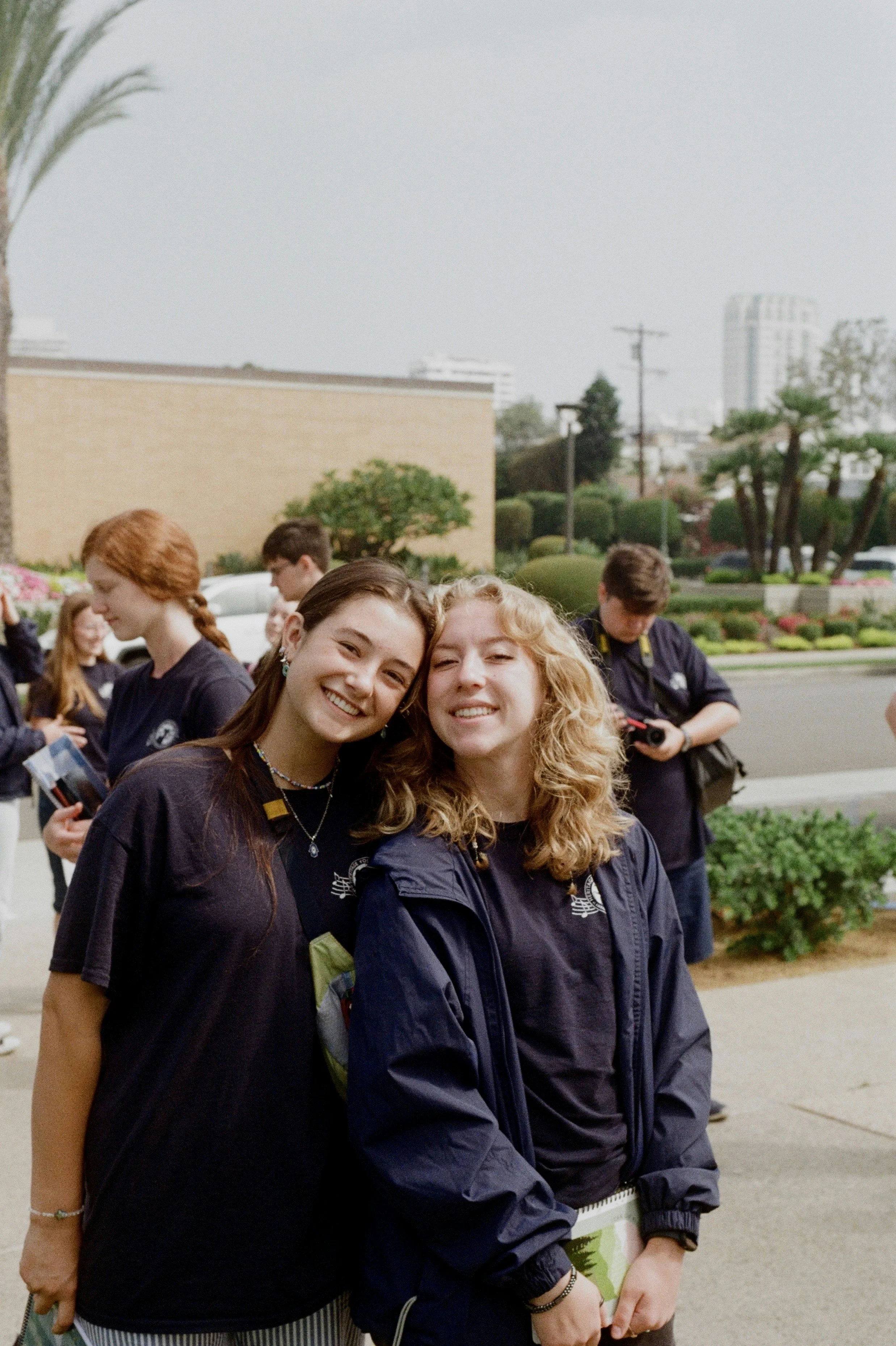Two smiling young women standing close together outdoors, surrounded by other people, with a green garden and buildings in the background.
