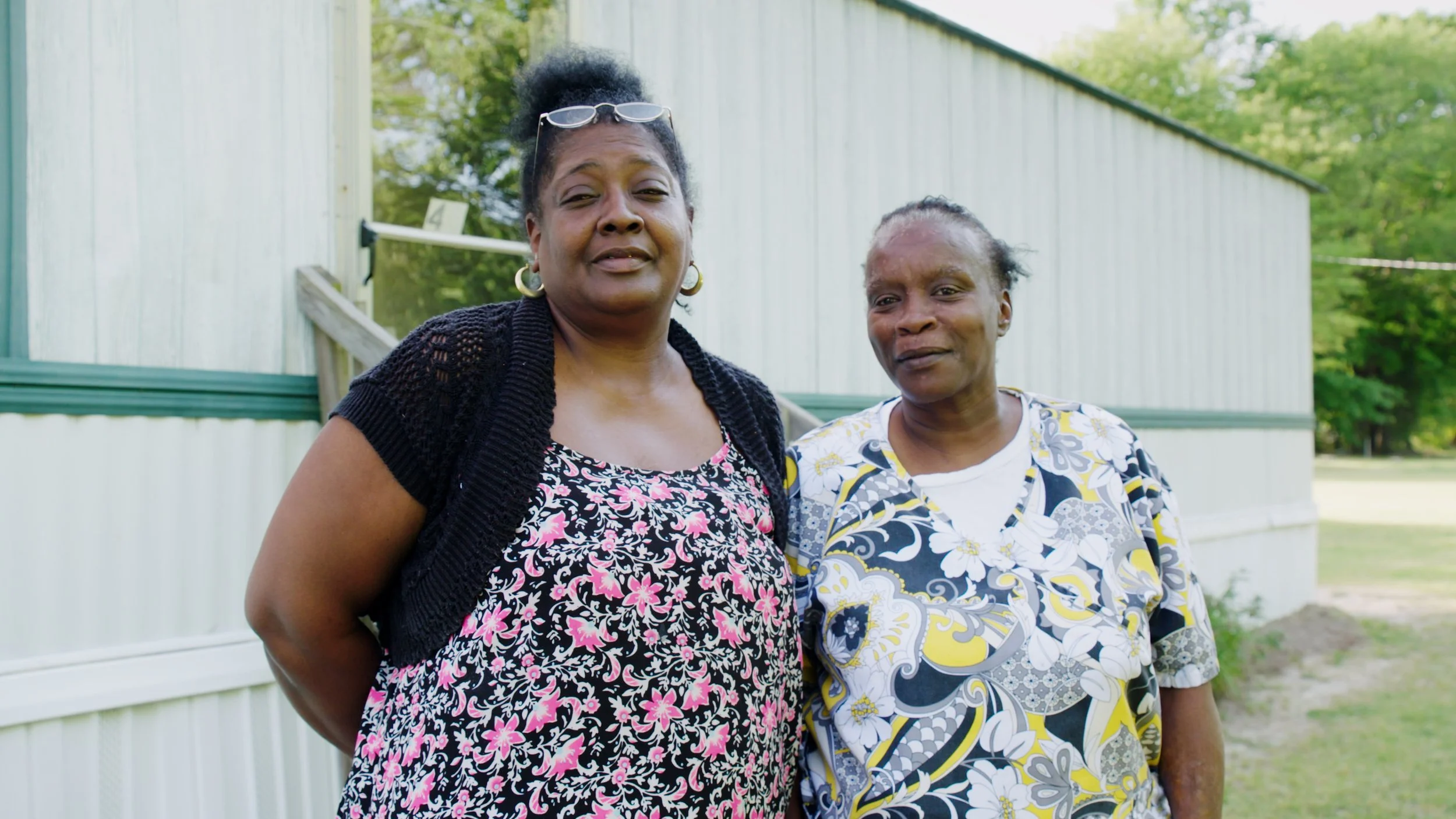 Two women impacted by the environmental impacts of hog farms in Eastern NC are standing outdoors in front of a white building with green trees in the background.