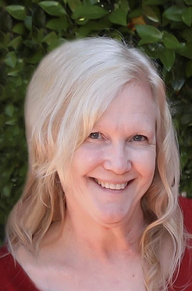 A woman with long, blonde, wavy hair smiling outdoors with green leafy background.