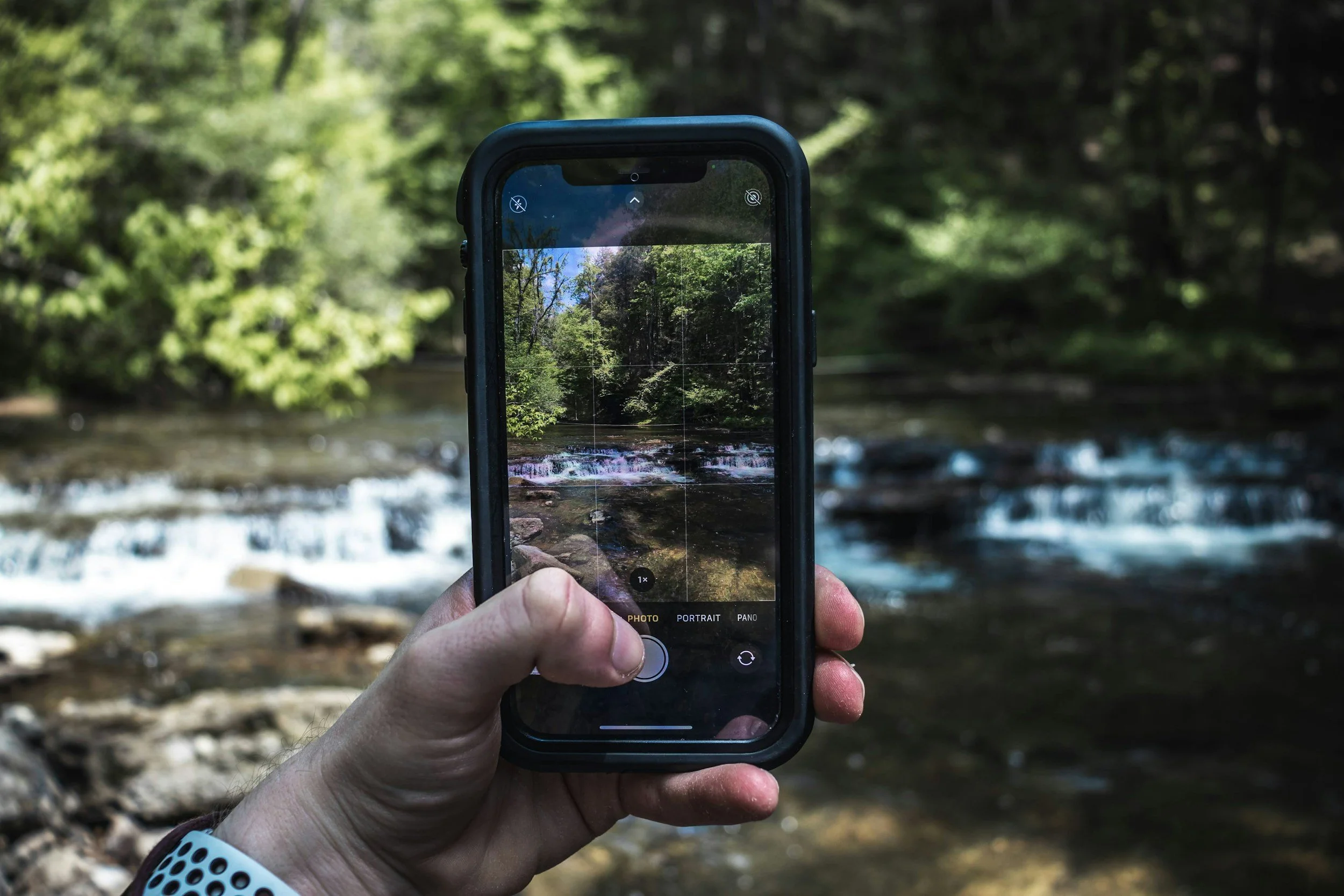 A person holding a smartphone taking a photo of a creek in a forested area.
