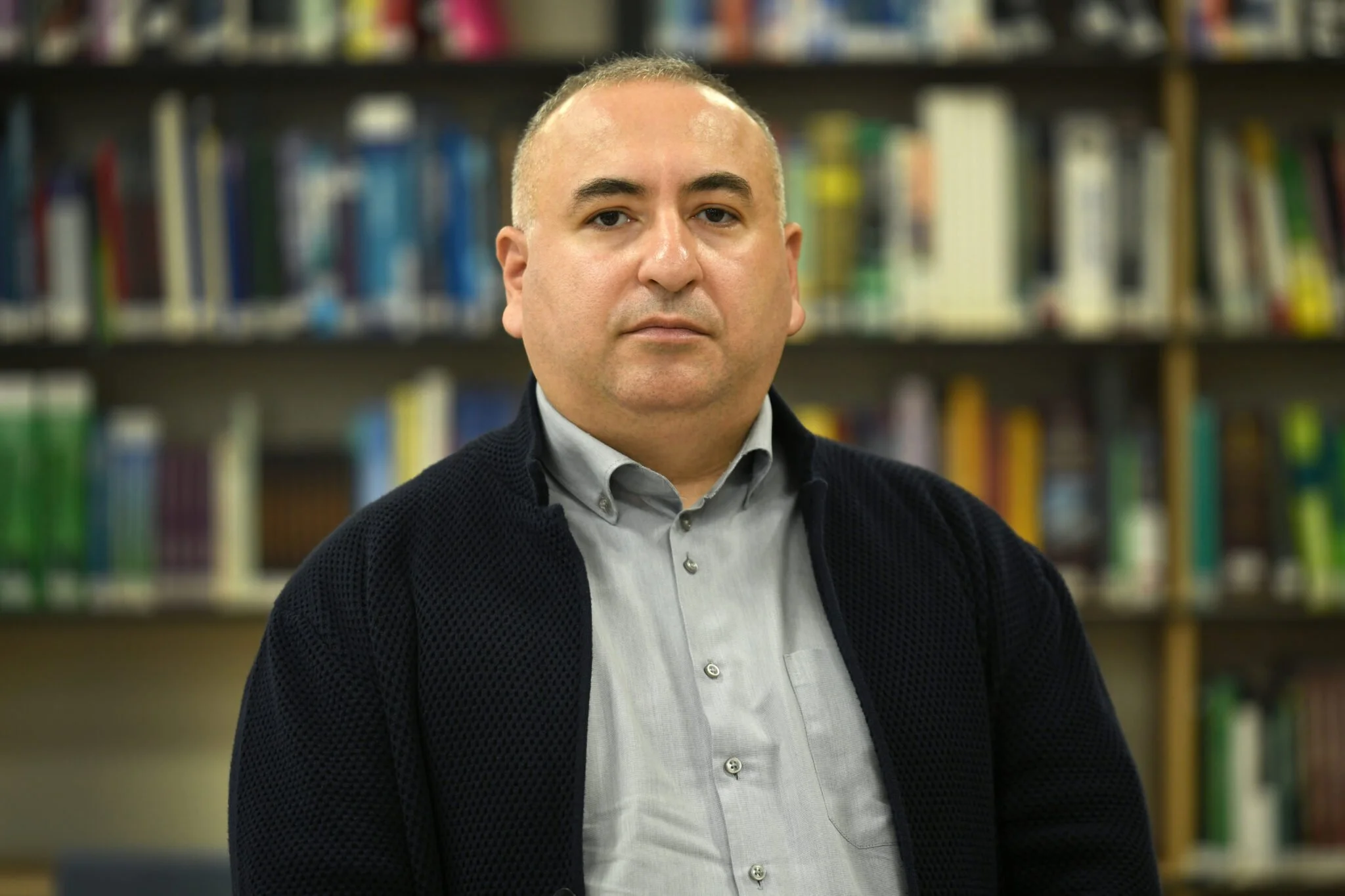 A man with a bald head and light skin, wearing a gray shirt and black sweater, stands in front of a bookshelf filled with colorful books.