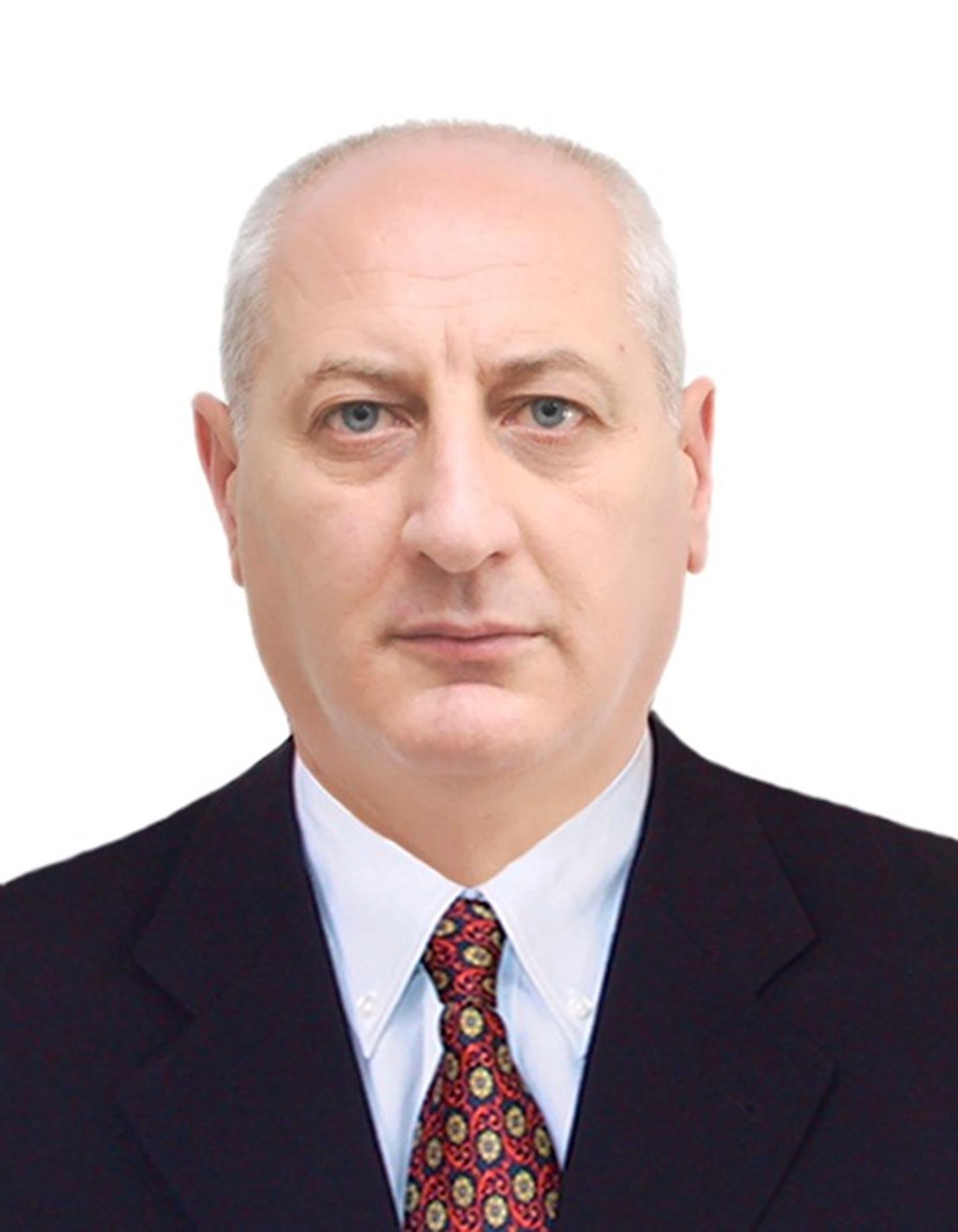 A middle-aged man with short, light-colored hair and blue eyes wearing a dark suit, white shirt, and a colorful patterned tie.