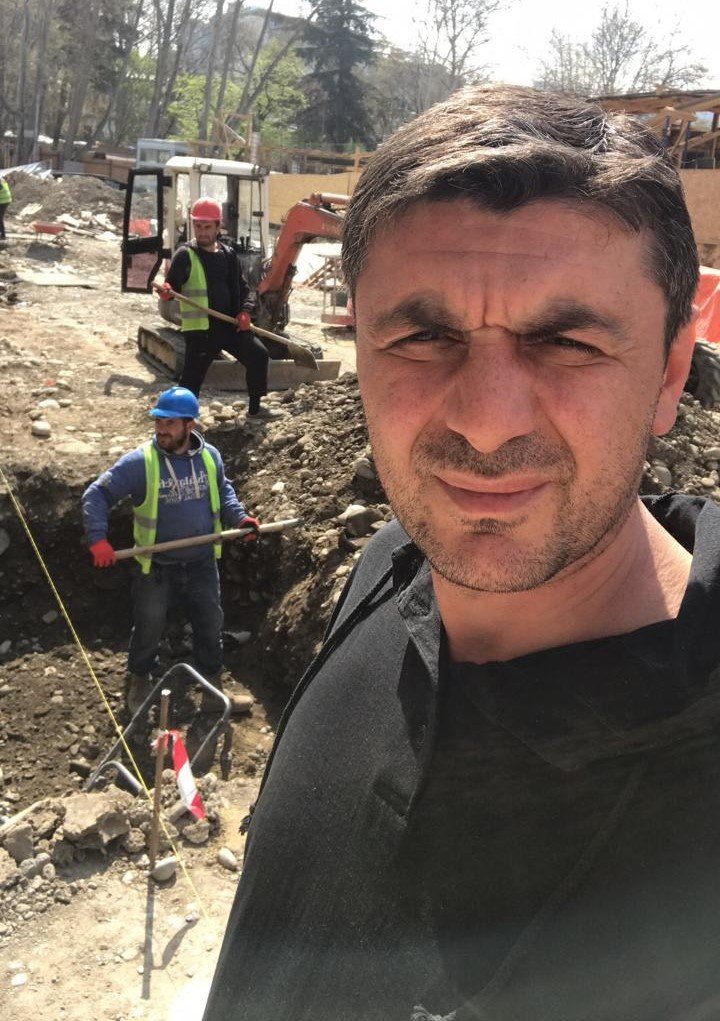 Man taking a selfie at a construction site with two workers digging in the background.
