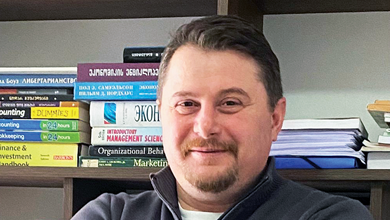 A man with short dark hair and a beard smiling in front of a bookshelf filled with books and papers.