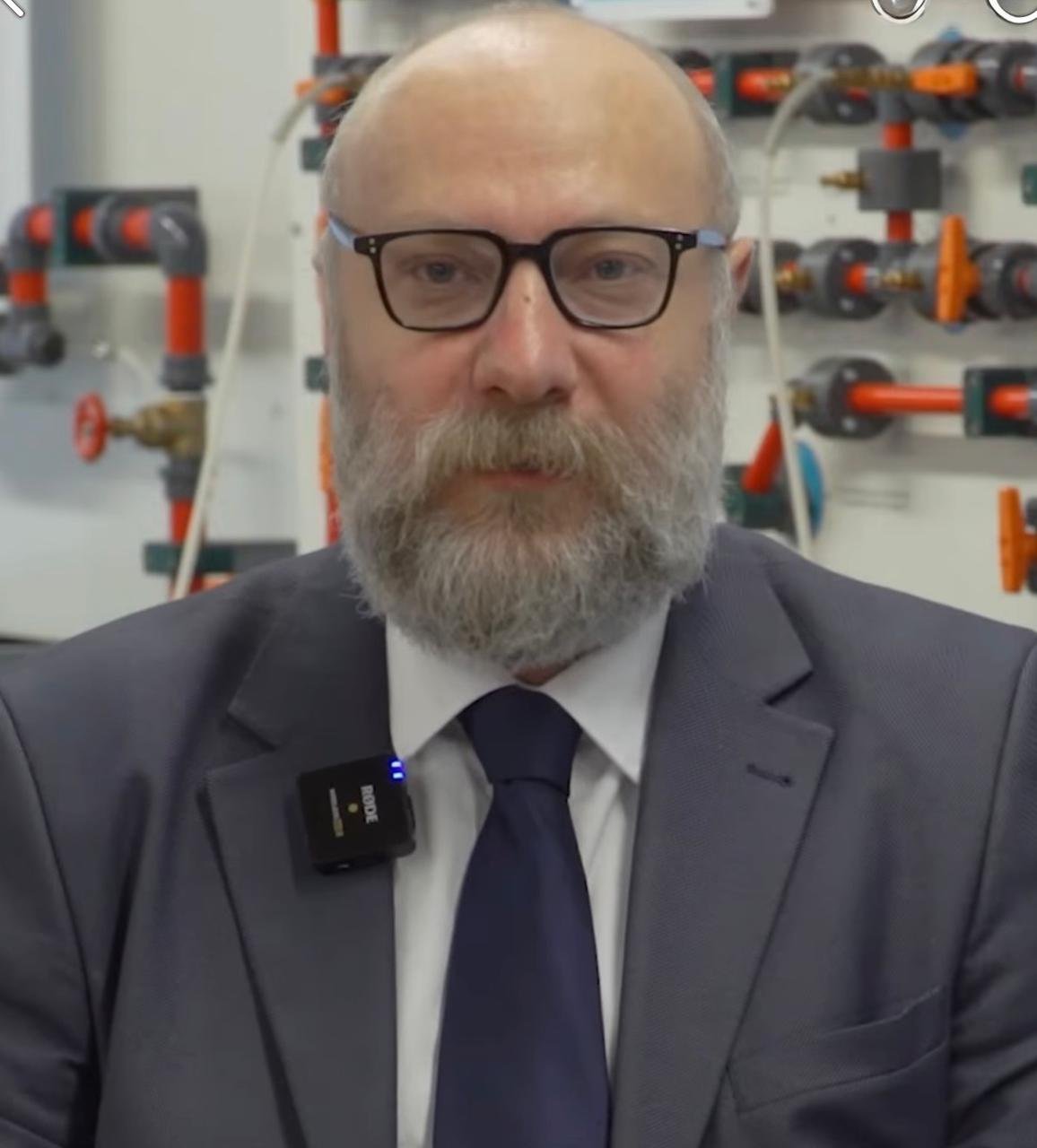 A middle-aged man with glasses and a beard wearing a suit and tie, standing in front of a wall of orange and gray plumbing pipes.