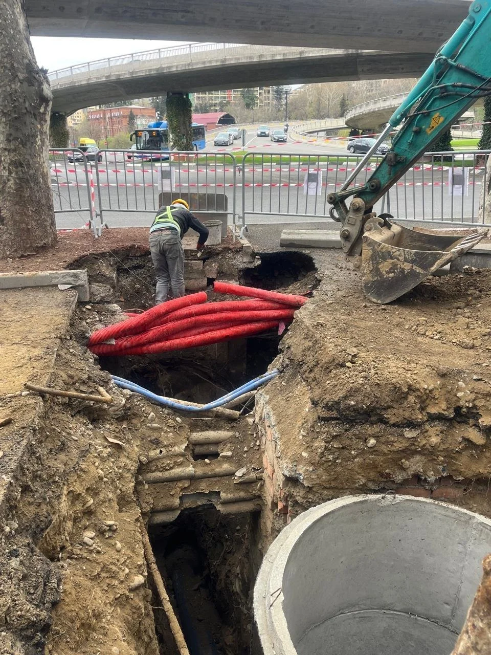 Construction worker in a reflective vest and helmet working in a large excavated hole with construction pipes and heavy machinery, under an overpass, with street and traffic in the background.