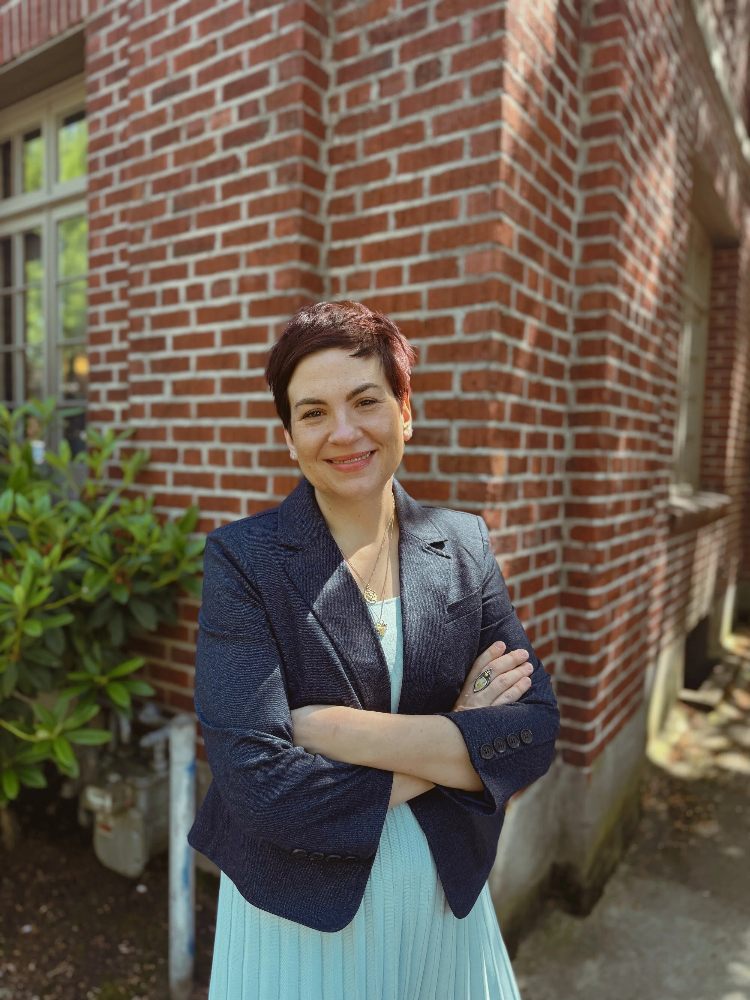 Co-founder and Managing Partner of The Whole Package LLC, Carrie Novak stands with her arms crossed in front of a red brick wall. Carrie is smiling and has brown hair and light skin tone. Carrie is wearing a navy blue jacket with light blue dress.