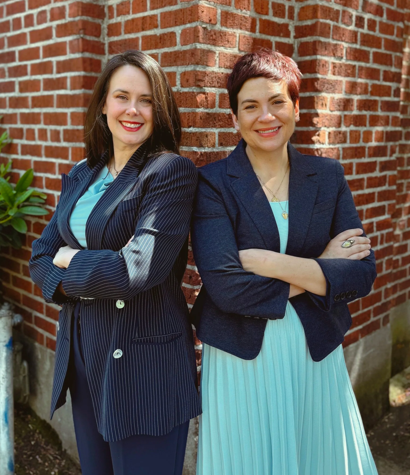 Co-founders of The Whole Package LLC, Carrie Novak and Maggie Clack, stand back to back in front of a red brick wall. The women both have brown hair and light skin tone, have their arms crossed, and are wearing blue jackets with light blue tops.