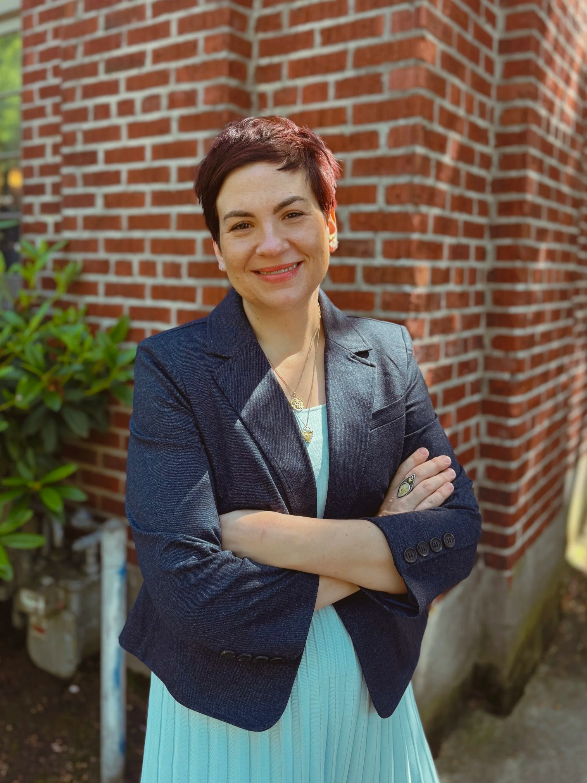 Co-founder and Managing Partner of The Whole Package LLC, Carrie Novak stands with her arms crossed in front of a red brick wall. Carrie is smiling and has brown hair and light skin tone. Carrie is wearing a navy blue jacket with light blue dress.