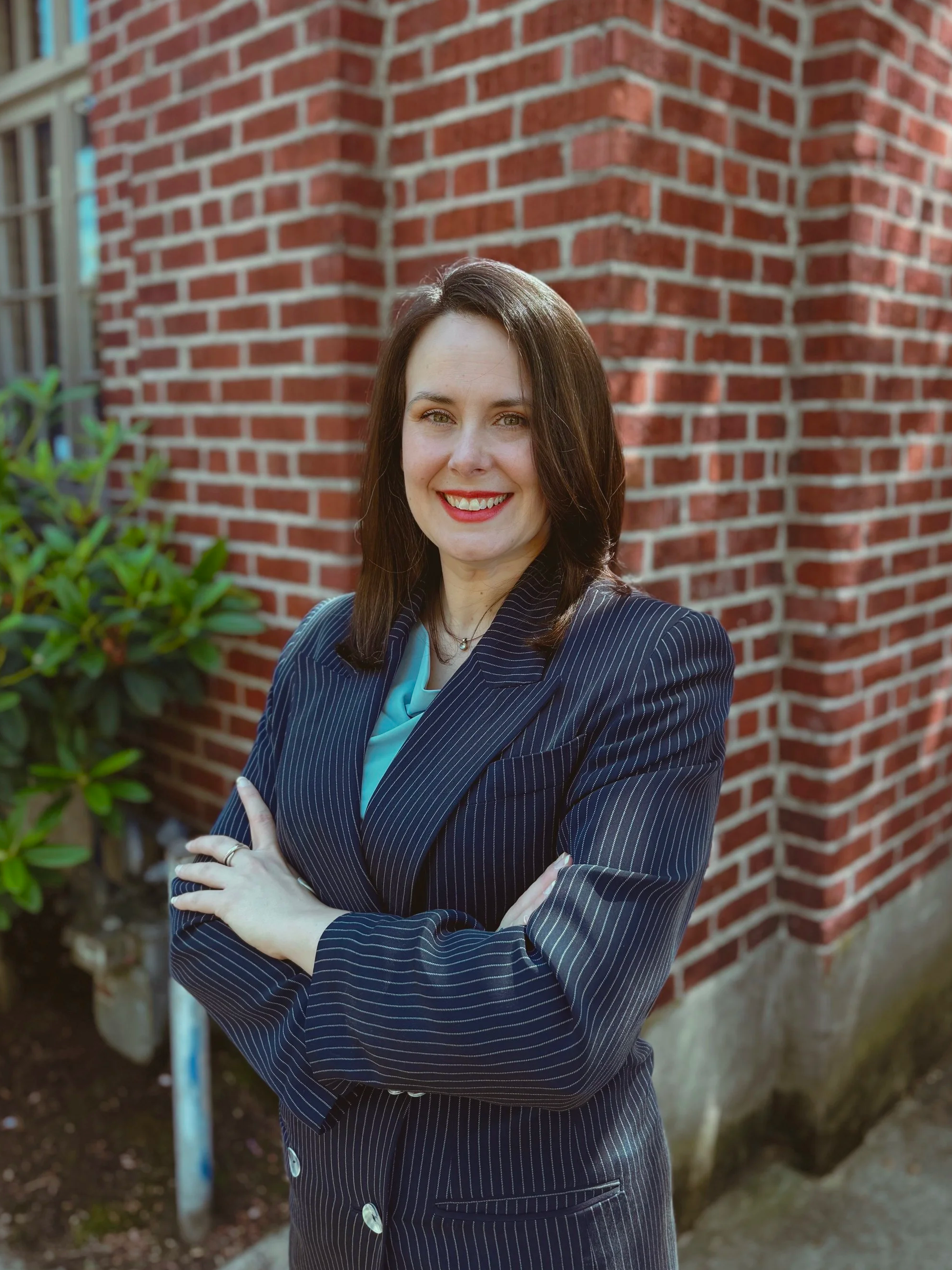 Co-founder and Managing Partner of The Whole Package LLC, Maggie Clack stands with her arms crossed in front of a red brick wall. Maggie is smiling, has brown hair and light skin tone. Maggie is wearing blue jacket, light blue top, and navy pants.