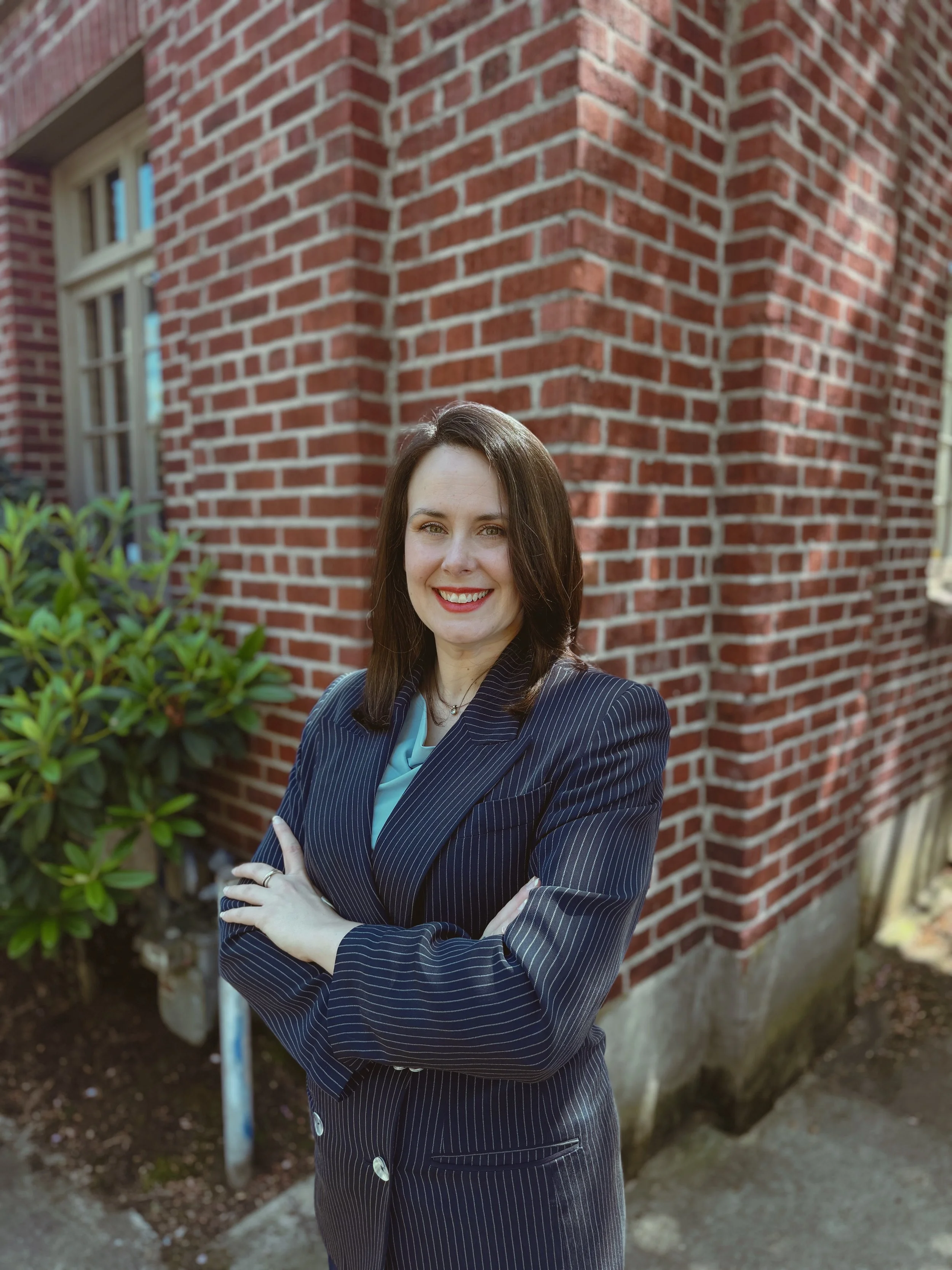 Co-founder and Managing Partner of The Whole Package LLC, Maggie Clack stands with her arms crossed in front of a red brick wall. Maggie is smiling, has brown hair and light skin tone. Maggie is wearing blue jacket, light blue top, and navy pants.