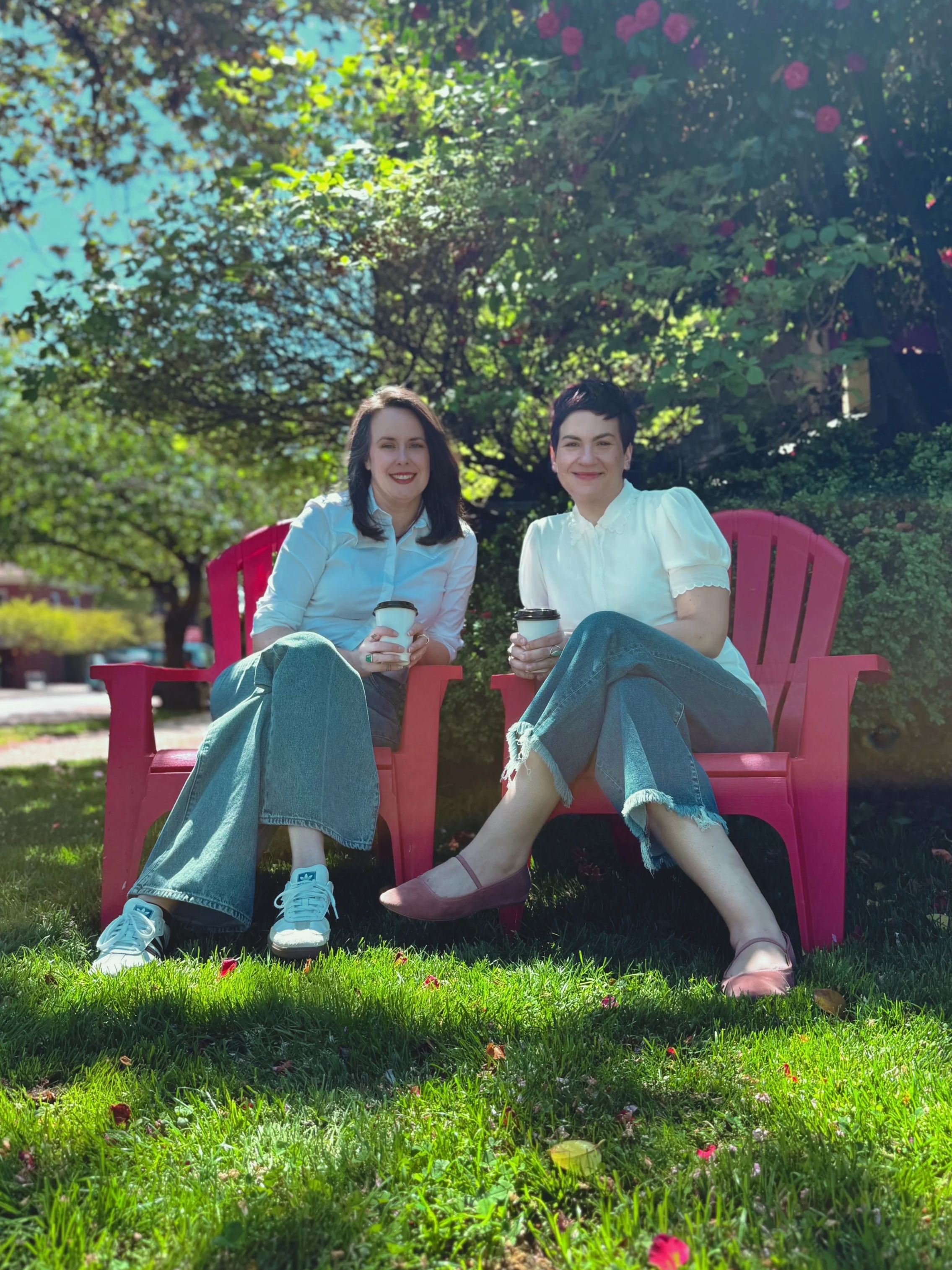 Co-founders of The Whole Package LLC, Carrie Novak and Maggie Clack, sit in red chairs on front of green foliage. The women both have brown hair and light skin tone, are holding a coffee cup, and are wearing white shirts and denim pants.