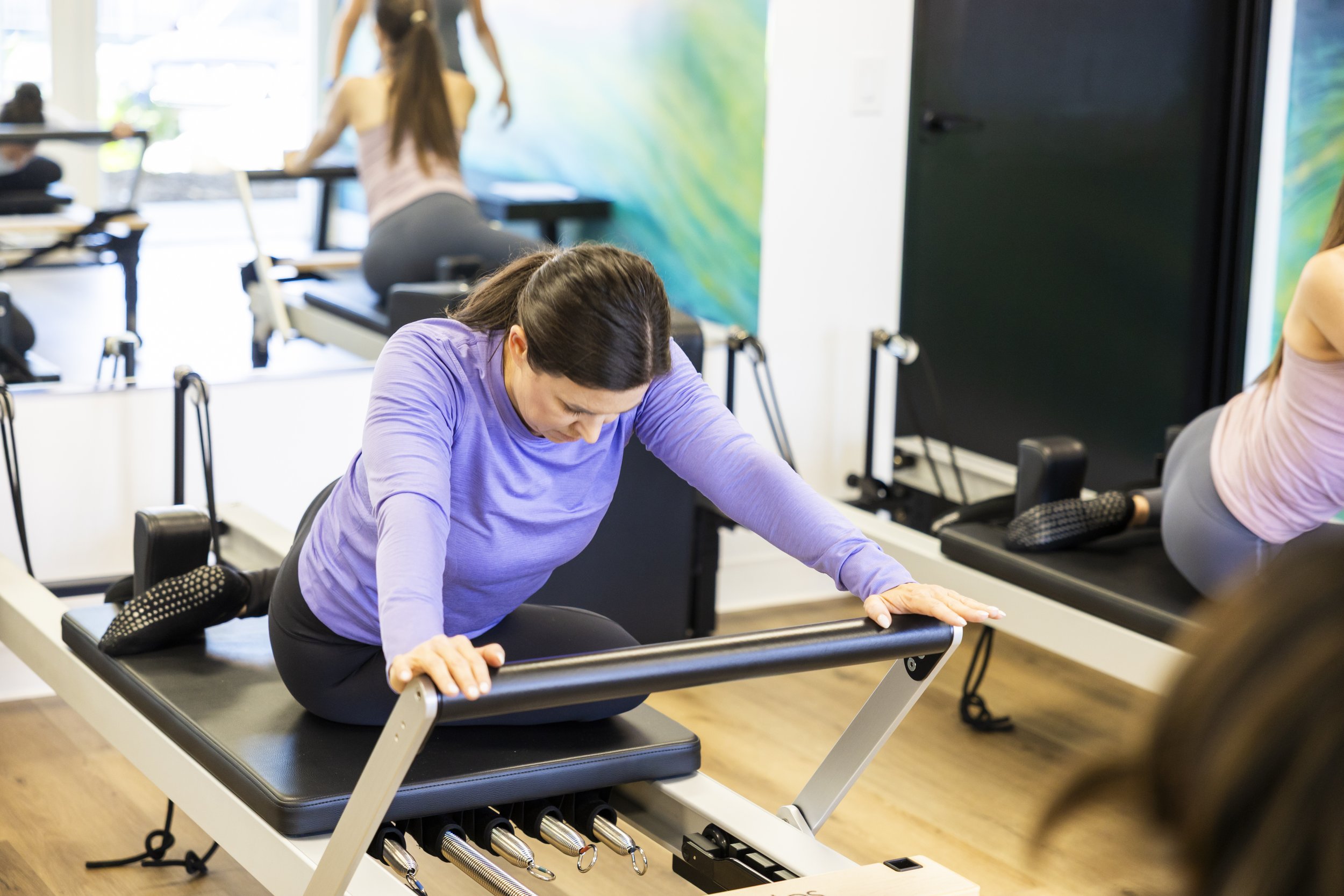 A woman in a purple shirt doing Pilates on a reformer machine at BOLD Pilates Encinitas with other women exercising nearby.