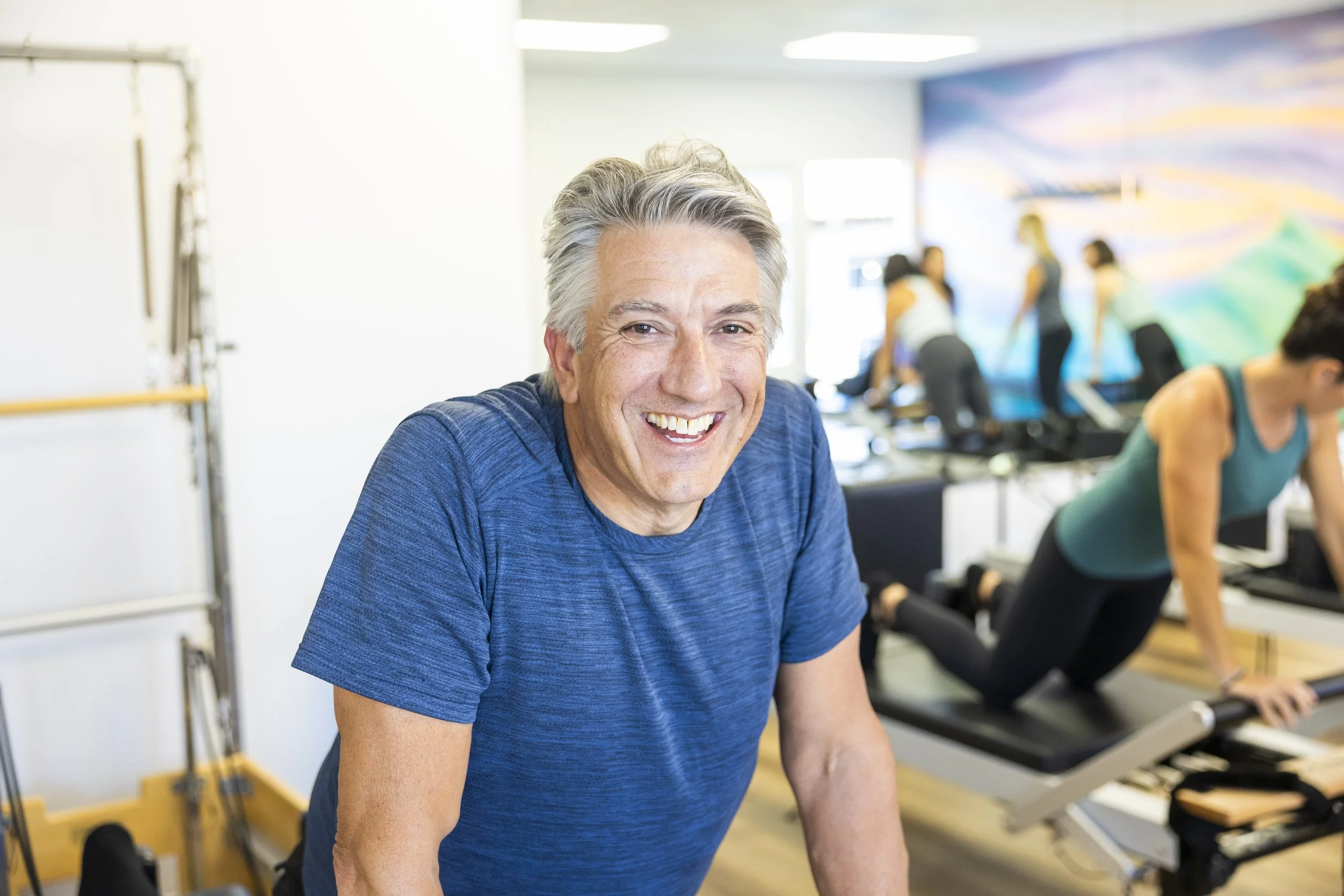 A smiling older man in a blue shirt at a fitness class, with people exercising in the background at BOLD Pilates in Encinitas.