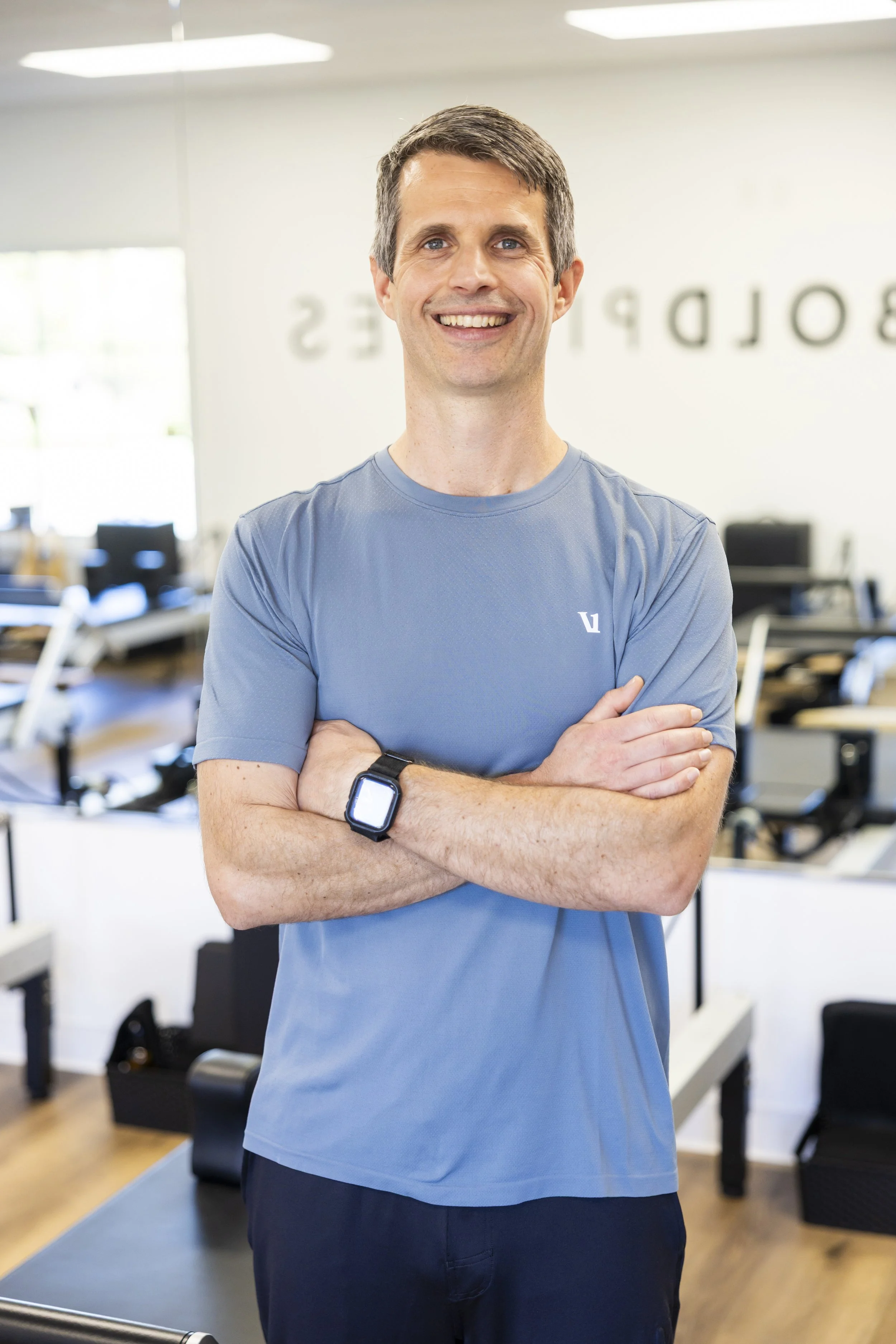 Smiling man with crossed arms wearing a blue athletic shirt and smartwatch, standing at BOLD Pilates Encinitas.