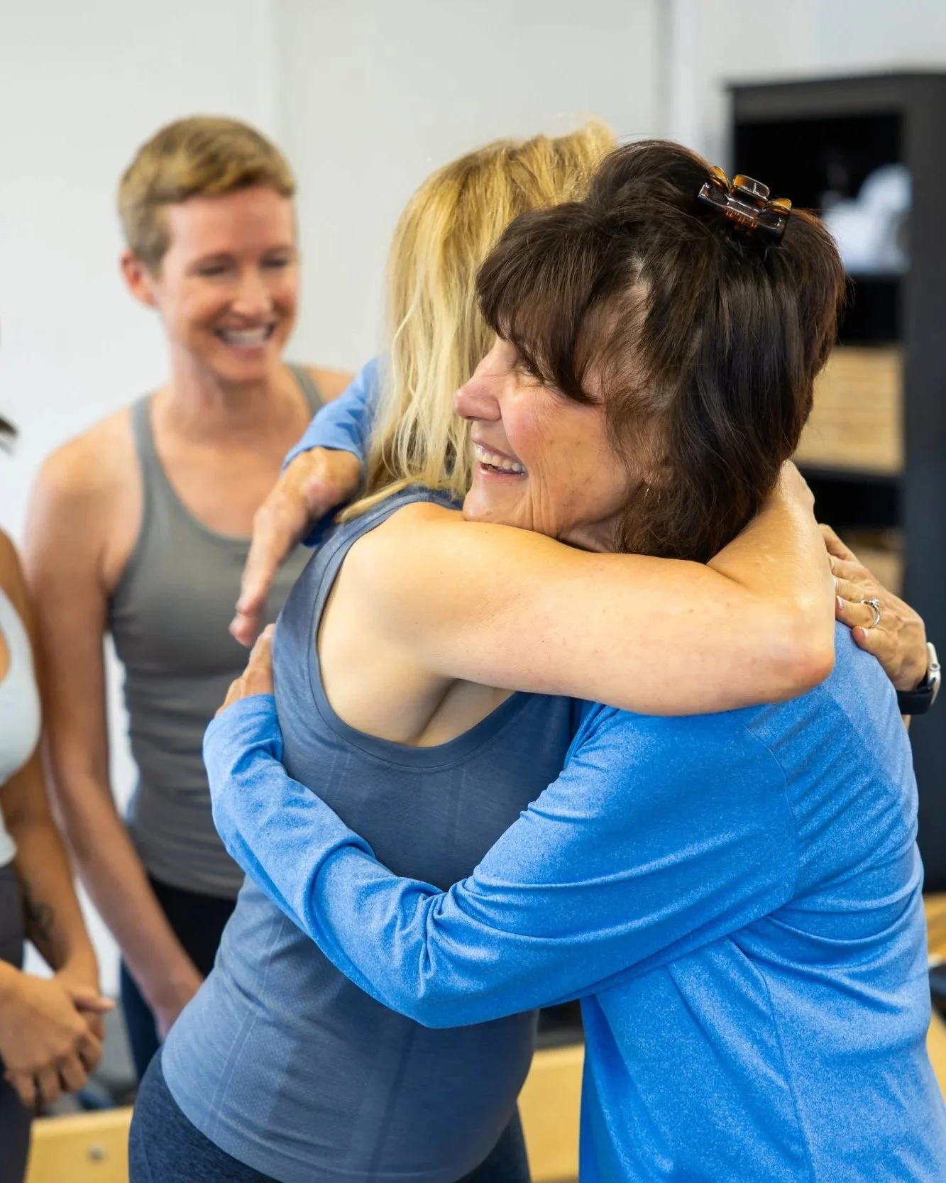Two women embrace in a hug, smiling happily, while two other women look on in the background, in an indoor setting.