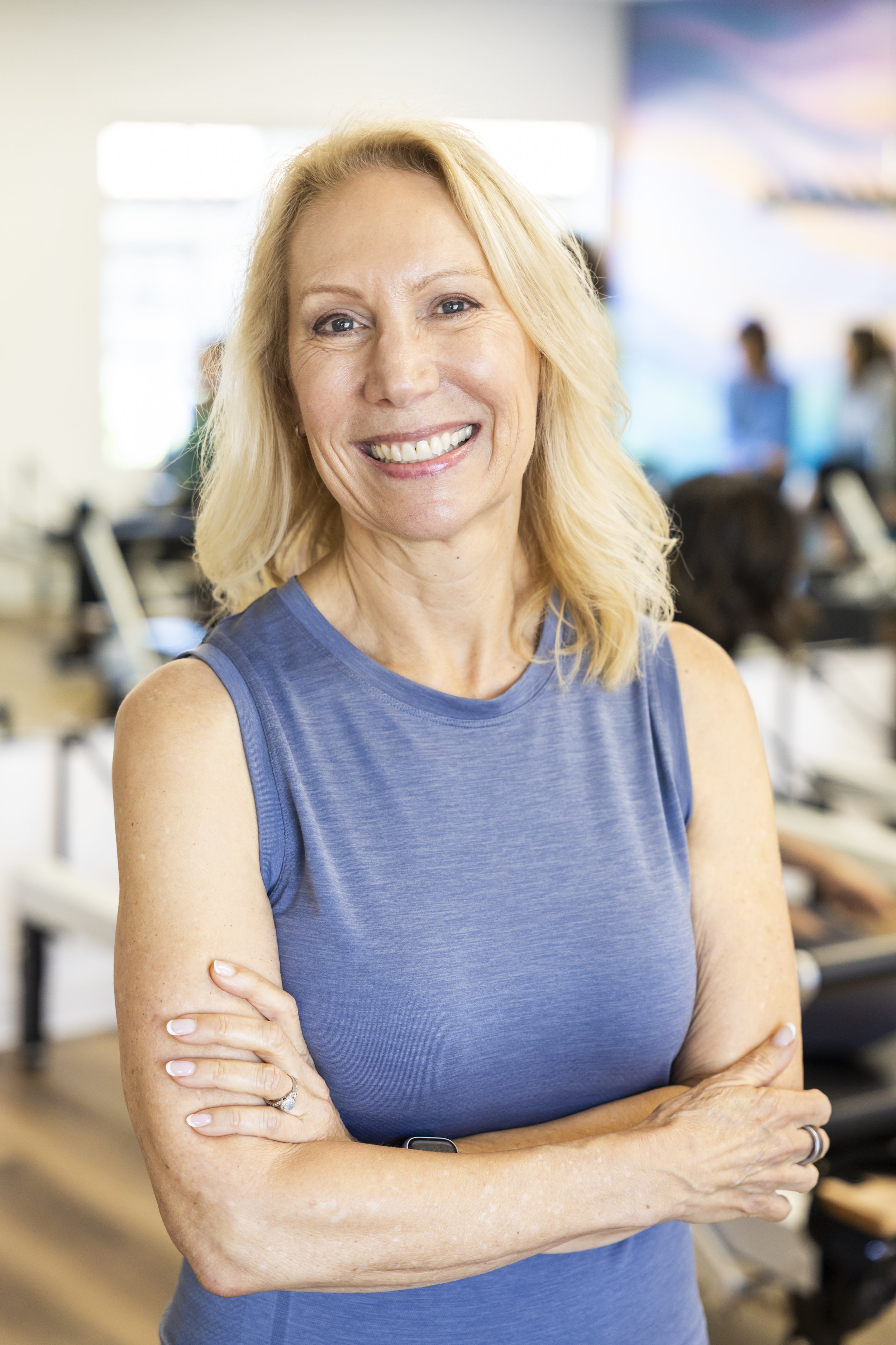 A middle-aged woman with blonde hair wearing a sleeveless blue top, smiling with crossed arms at BOLD Pilates Encinitas with exercise equipment and blurred individuals in the background.