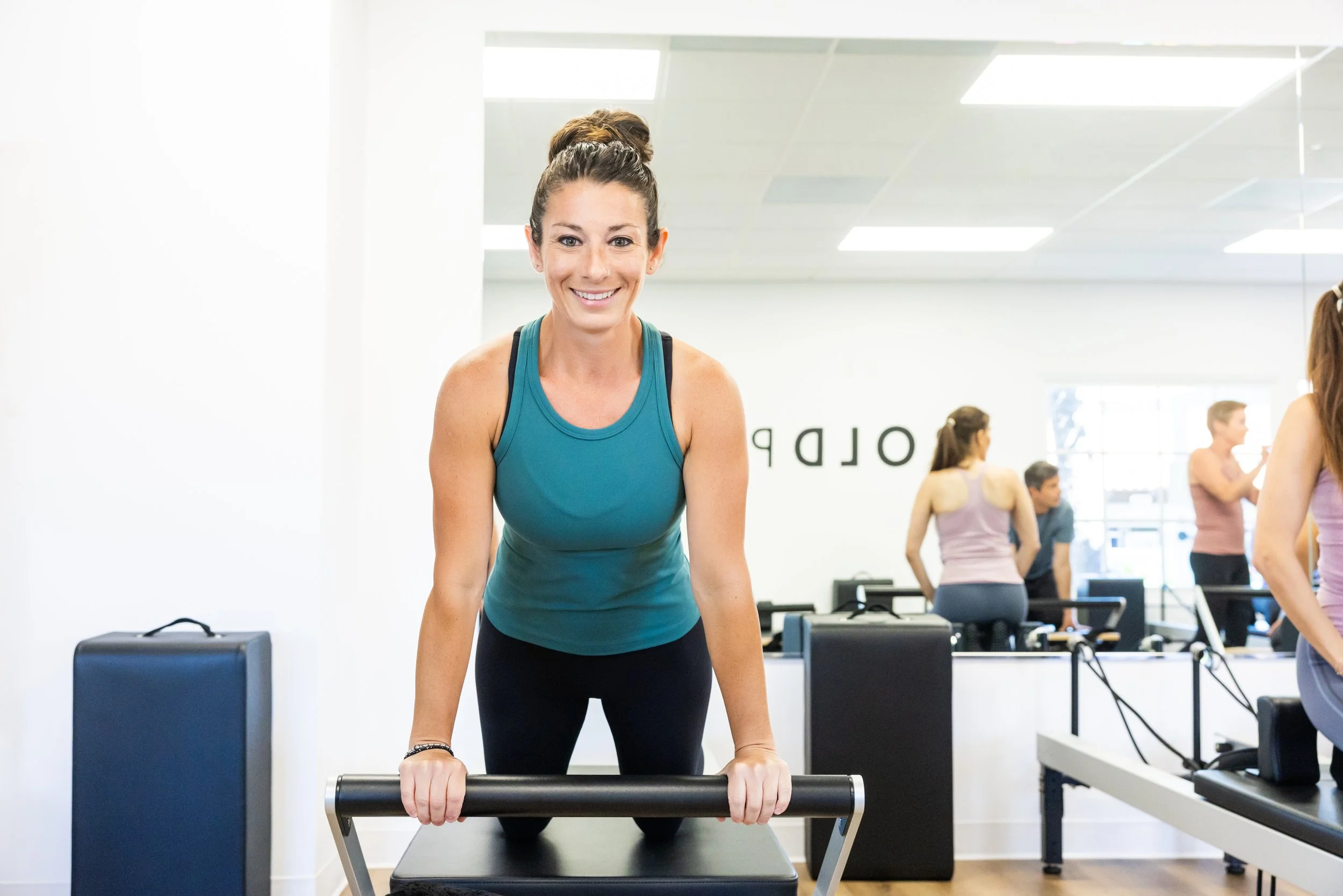 A woman in athletic clothing doing a push-up on a Pilates reformer at BOLD Pilates Encinitas, with other participants and exercise equipment visible in the background.
