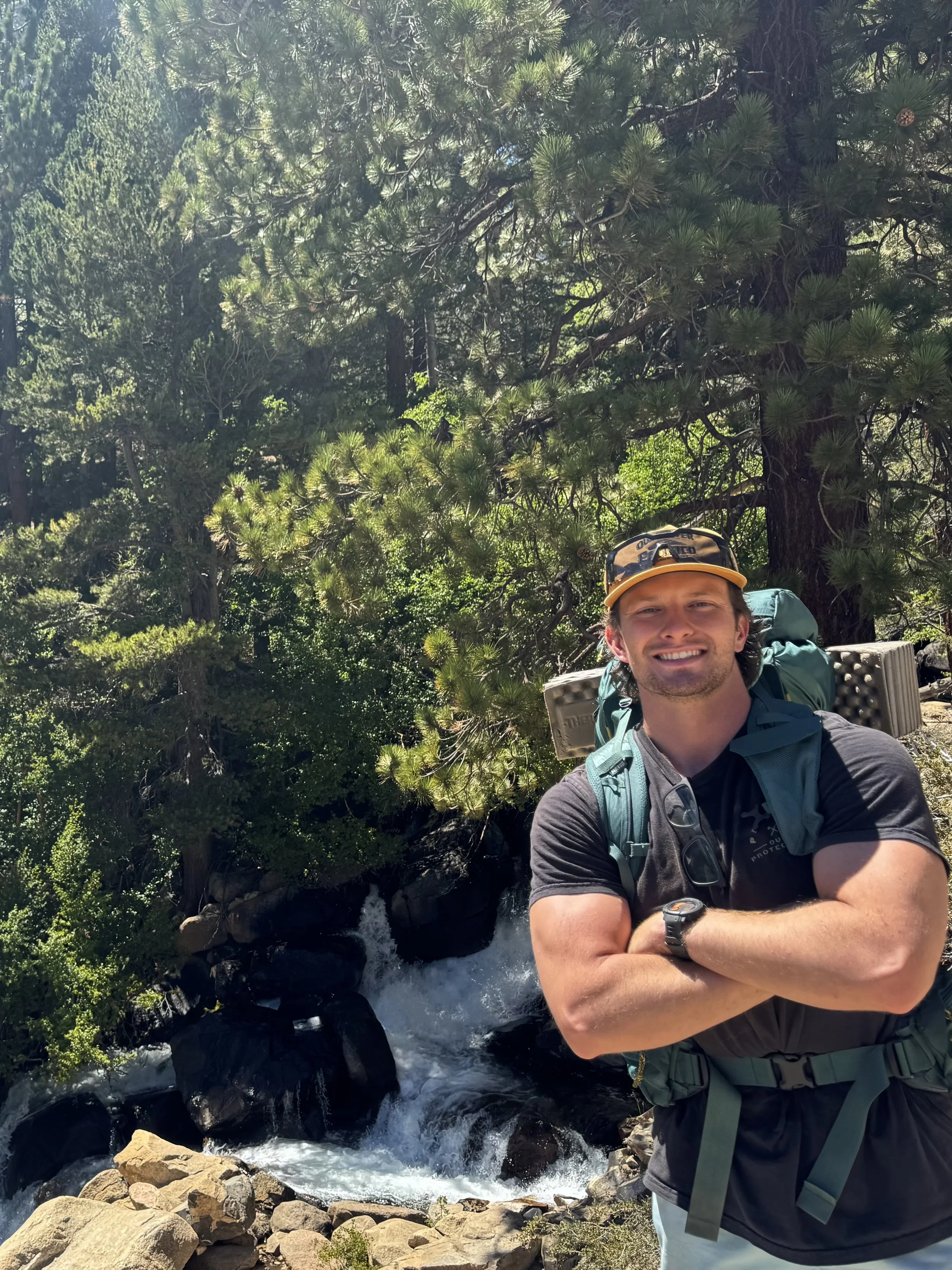 Smiling man with a backpack and wristwatch standing in front of a waterfall in a forested area.