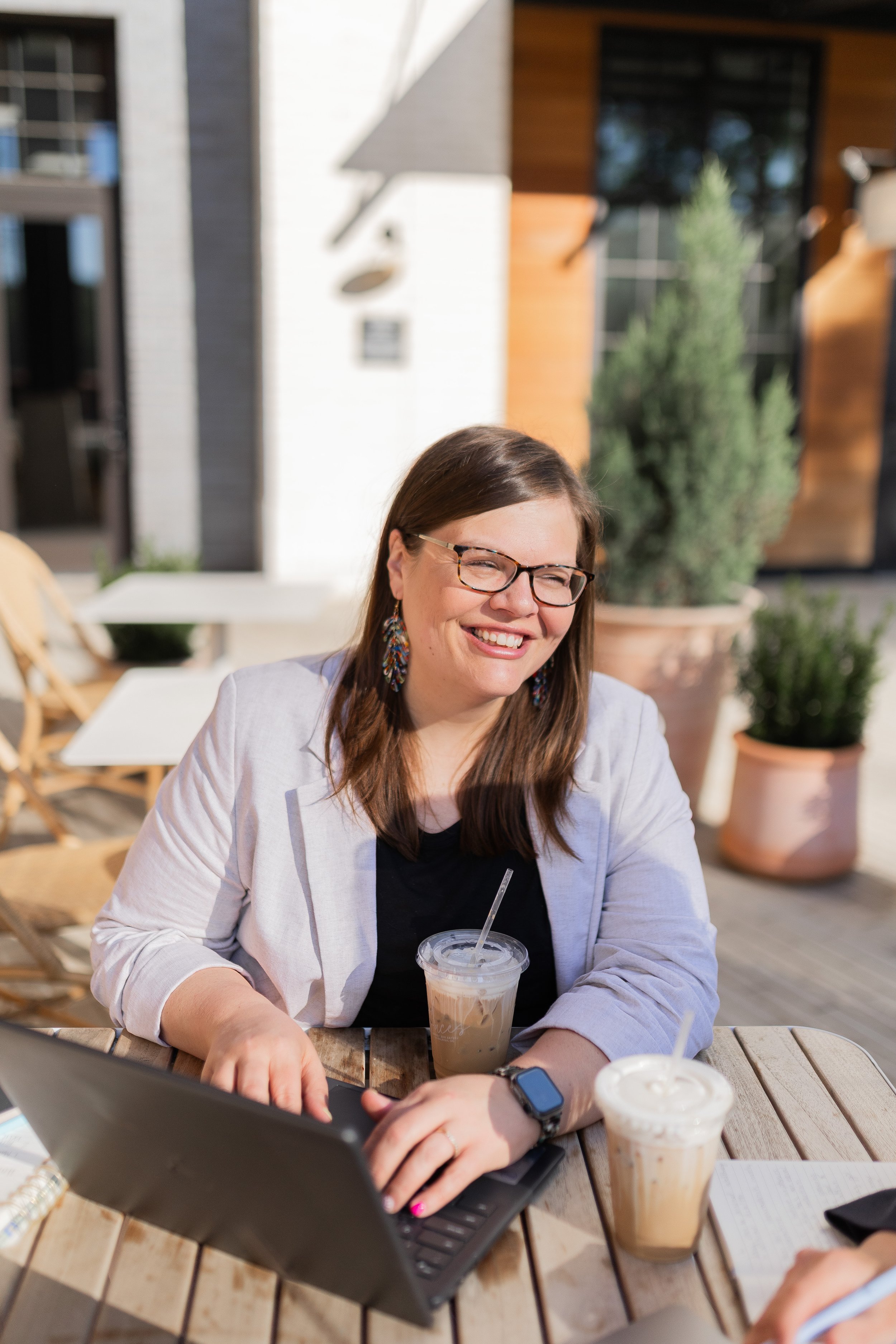 Smiling woman with glasses and long brown hair leaning against a wooden wall, outdoors.