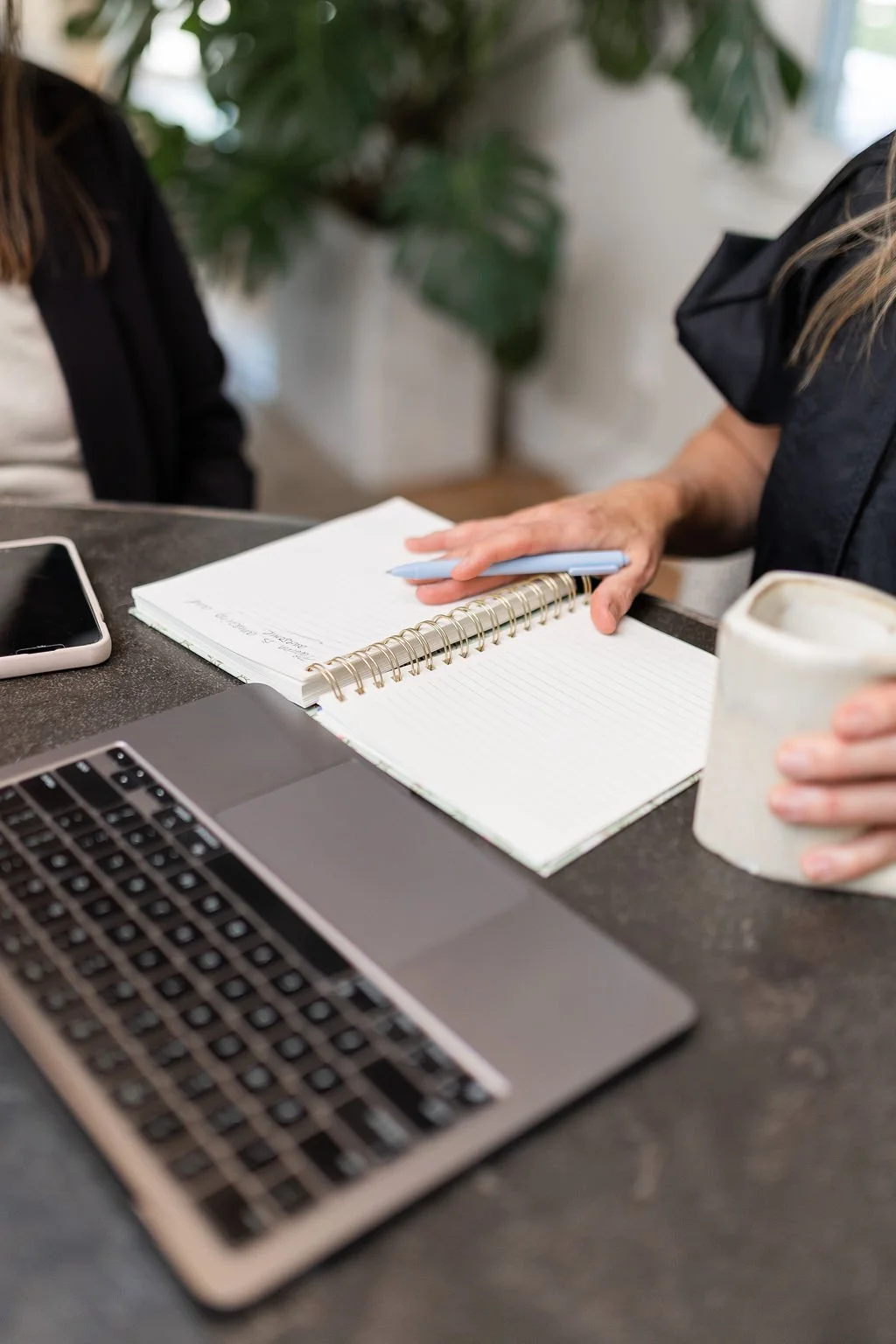 Close-up of a workspace with a laptop, smartphone, notebook, pen, and a mug on a dark desk, with two people sitting across.
