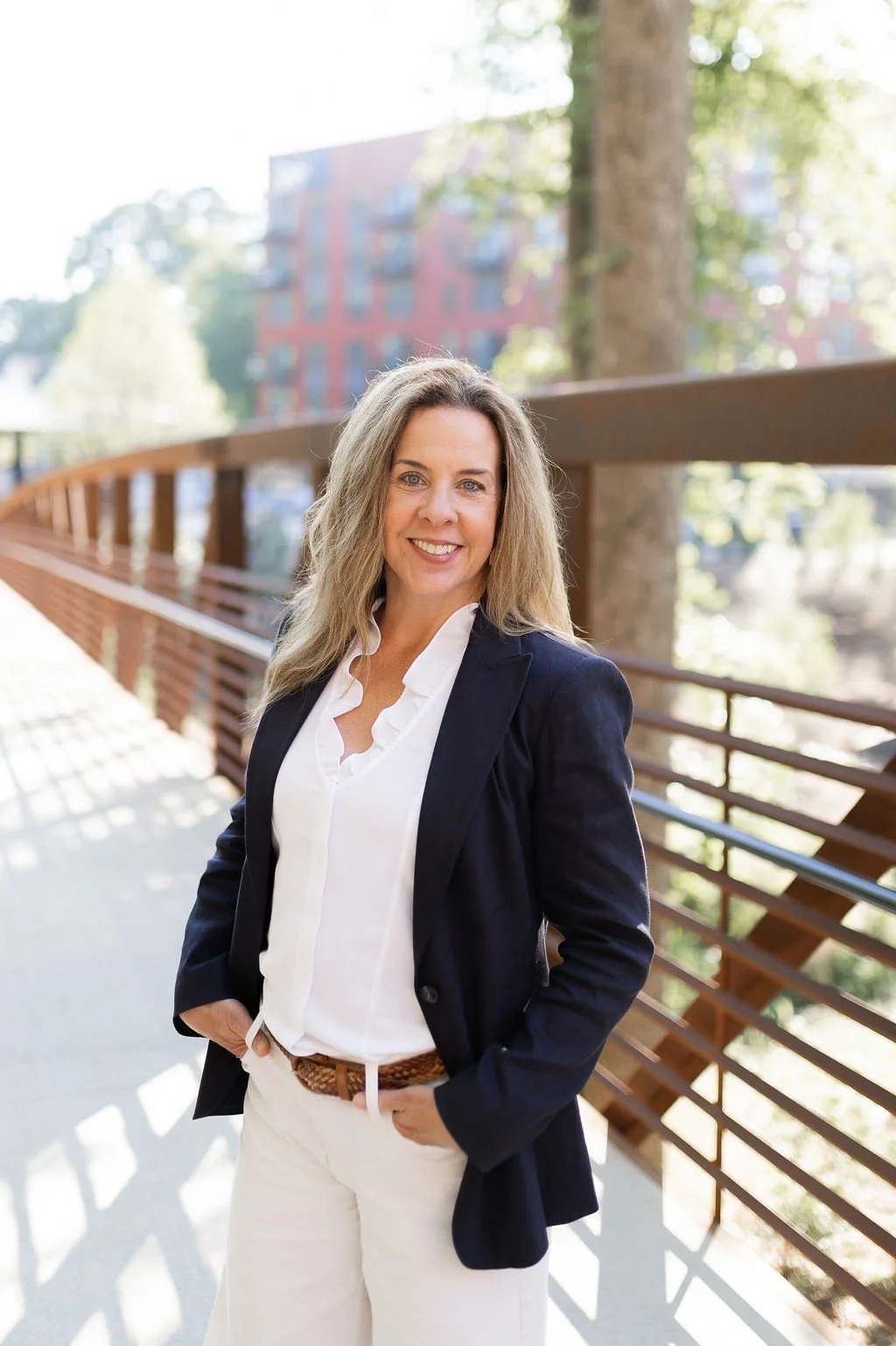 A woman with long blonde hair wearing a white blouse, navy blazer, and cream pants, smiling on an outdoor bridge with trees and a building in the background.