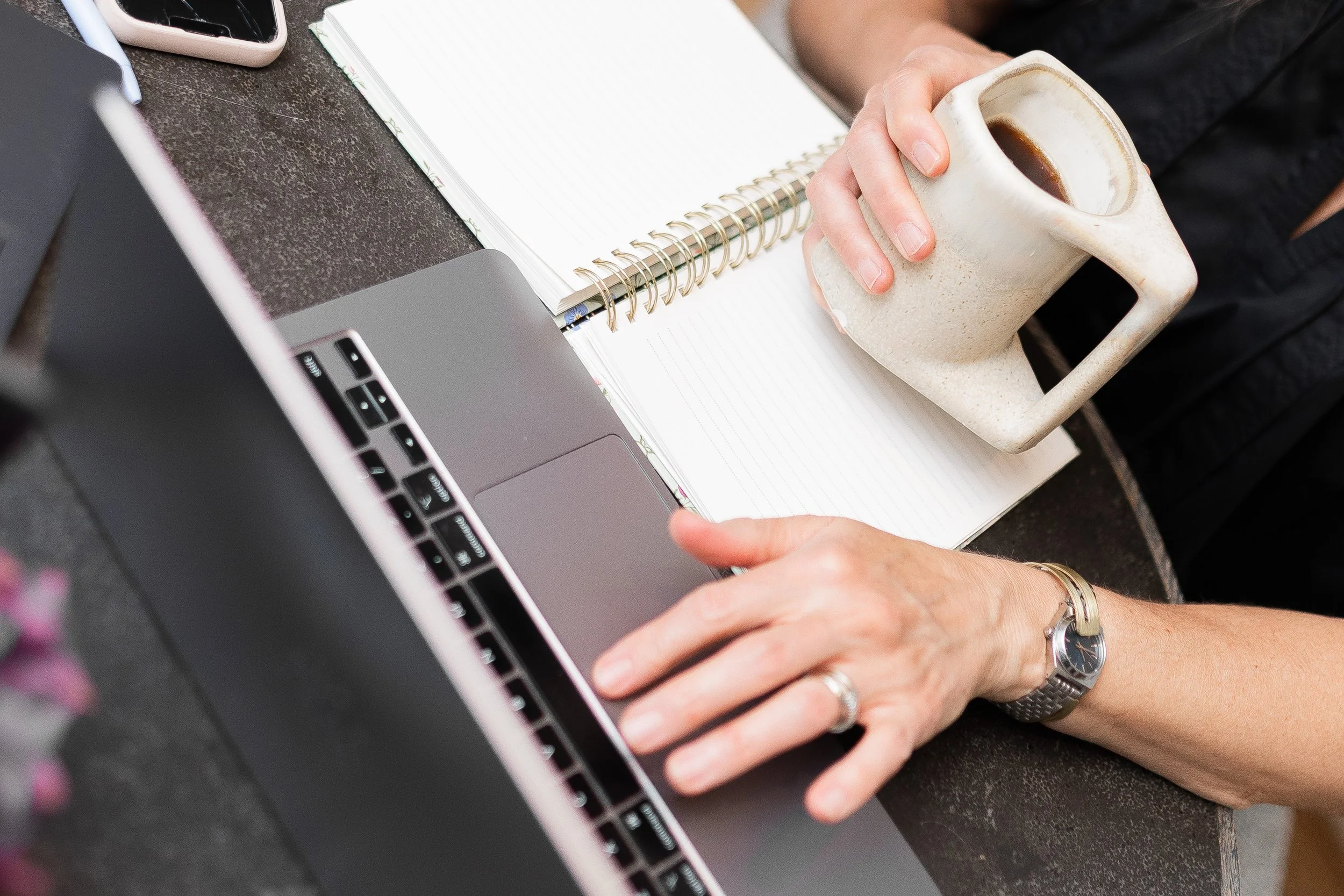 A person holding a coffee mug while sitting at a desk with a laptop, a notebook, and a smartphone.