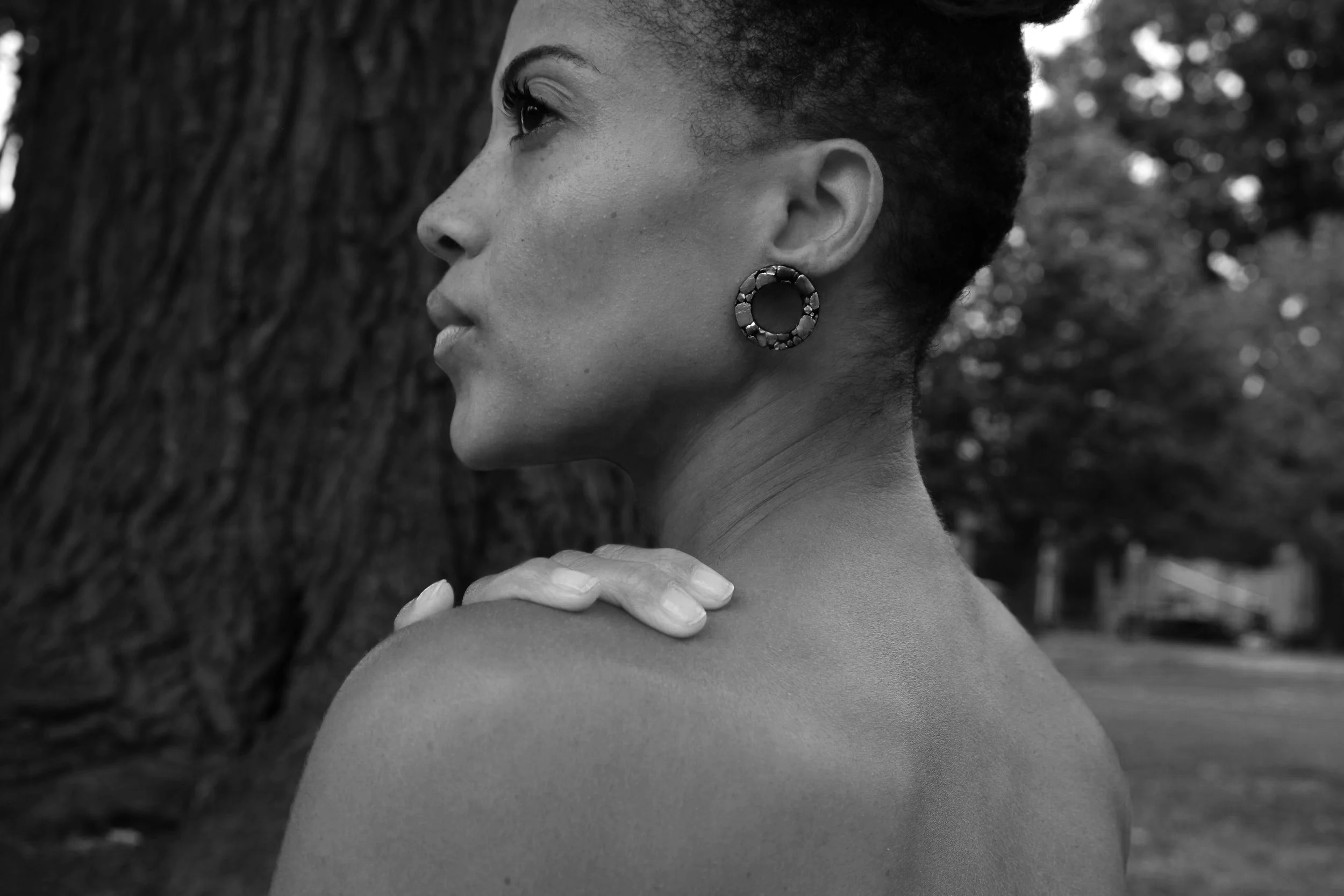 Black and white close-up of a woman with short curly hair, wearing a large circular earring, looking to the side with her hand resting on her shoulder, standing outdoors near a tree.