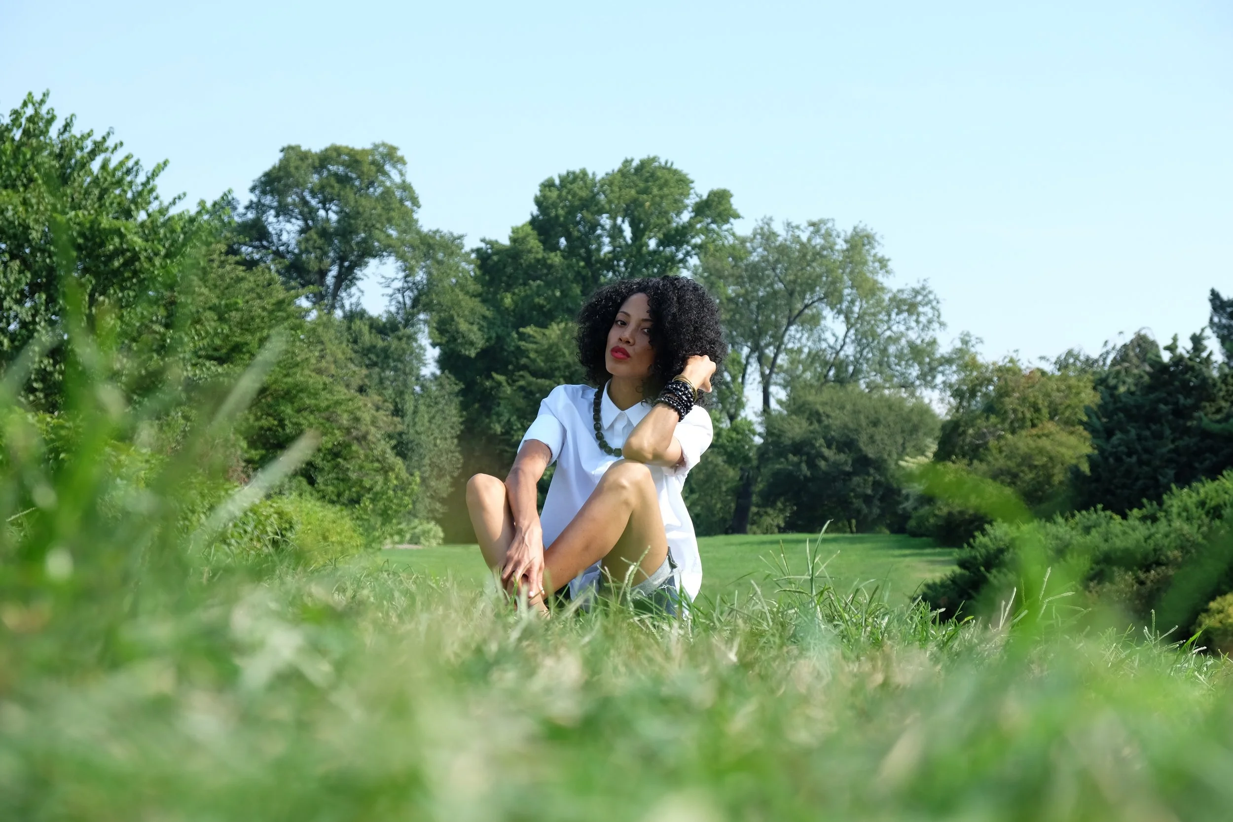 A woman with curly black hair and red lipstick sitting outdoors on grass among trees, wearing a white shirt and dark jewelry.