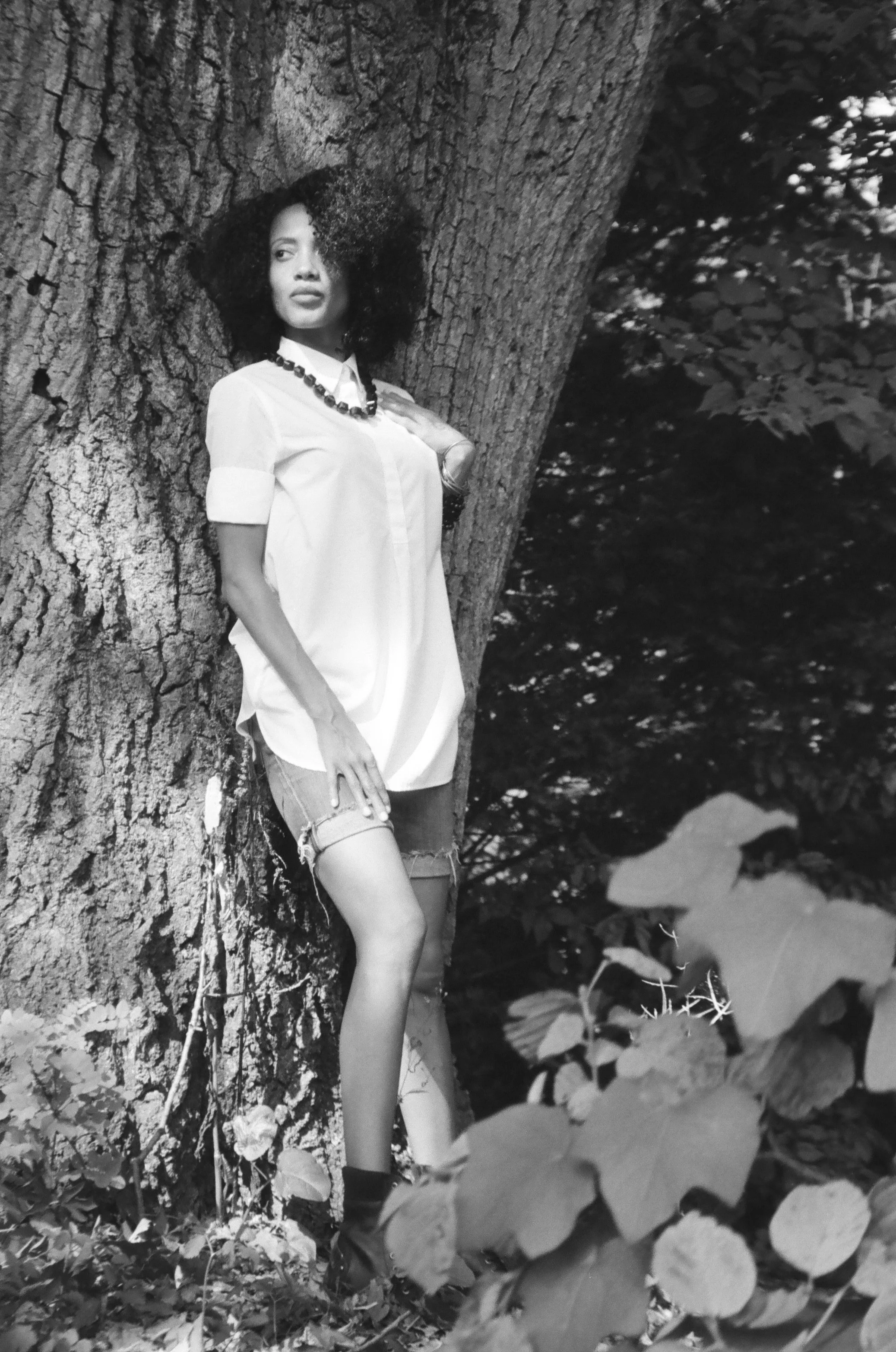 A woman with curly hair stands beside a large tree in a shaded outdoor setting, wearing a light-colored shirt, shorts, and boots, with leaves and water visible in the background.