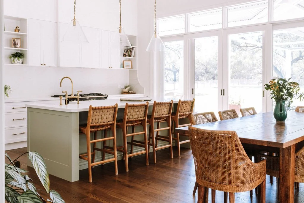 Bright, airy kitchen with white cabinets, a kitchen island, five barstools with woven seats, a dining table with rattan chairs, a vase with flowers, and large windows letting in natural light.
