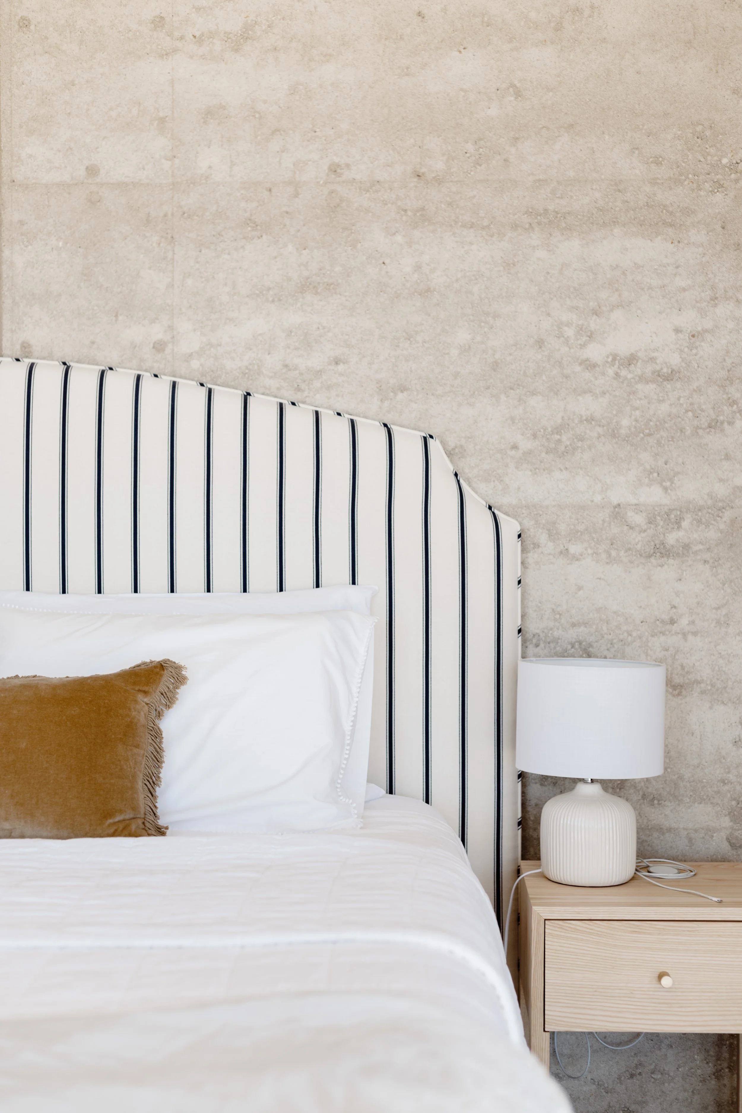Part of a neatly made bed with white sheets, a white pillow, and a brown pillow on top, next to a light wooden nightstand with a white lamp, against a textured beige wall with a striped headboard.