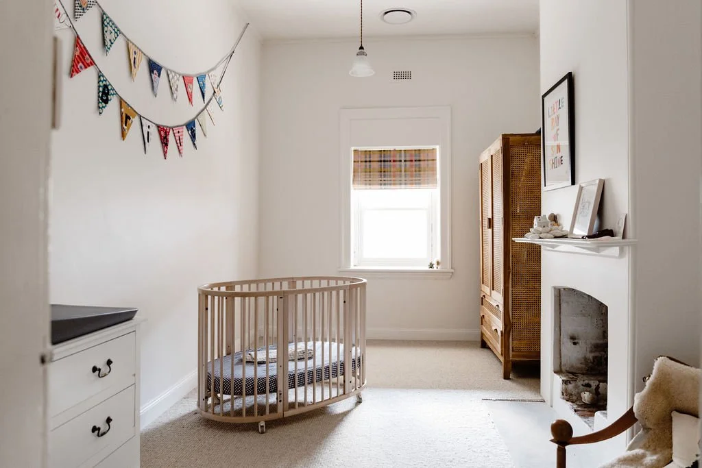 A cozy nursery with a wooden crib, a white dresser, a small window with a plaid curtain, and a decorative banner on the wall. There is a fireplace with decorations on the mantle and a woven cabinet in the corner.