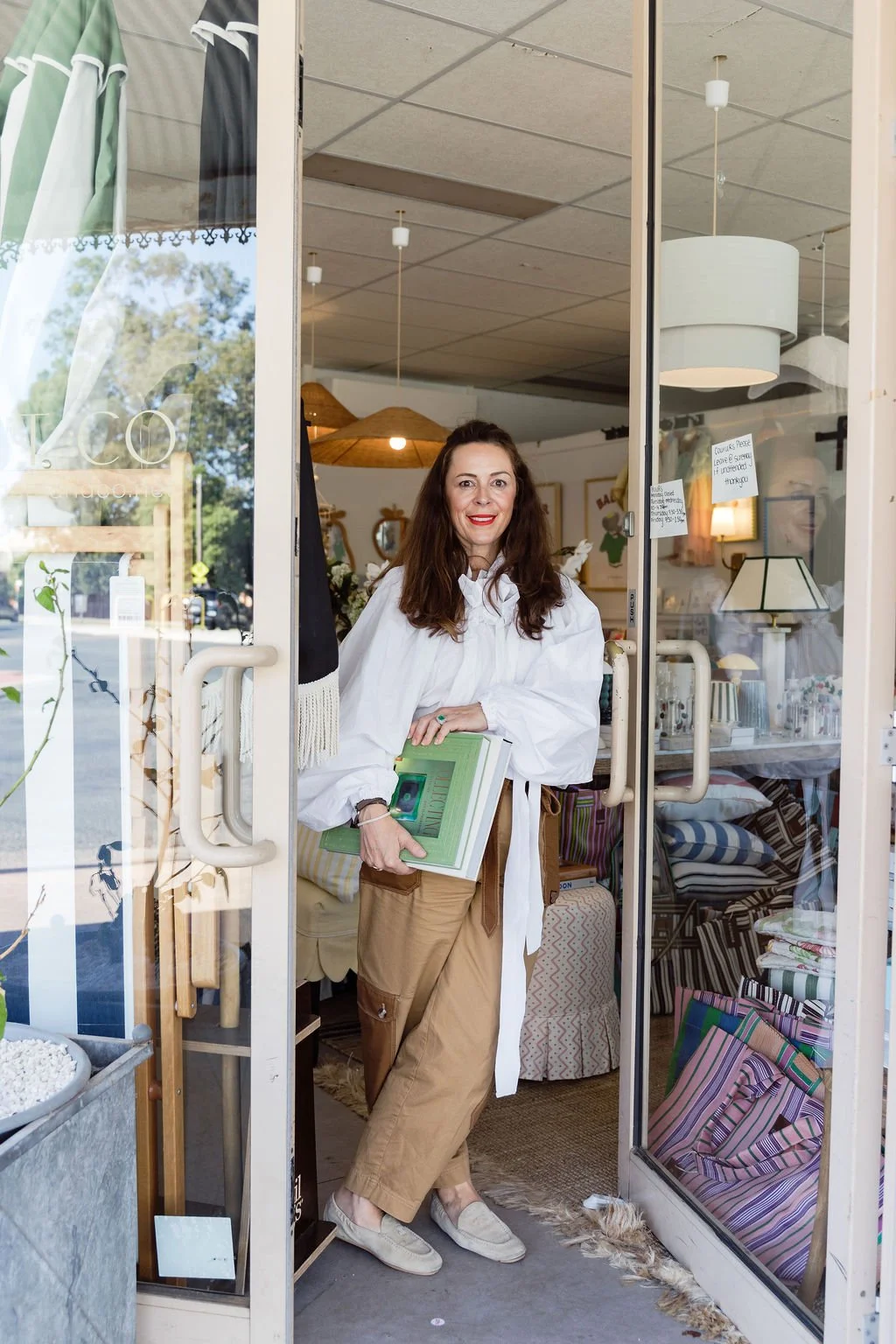 A woman with long brown hair, wearing a white shirt and beige pants, stands inside a store, holding a green book, smiling at the camera, with various home decor items visible inside the shop.