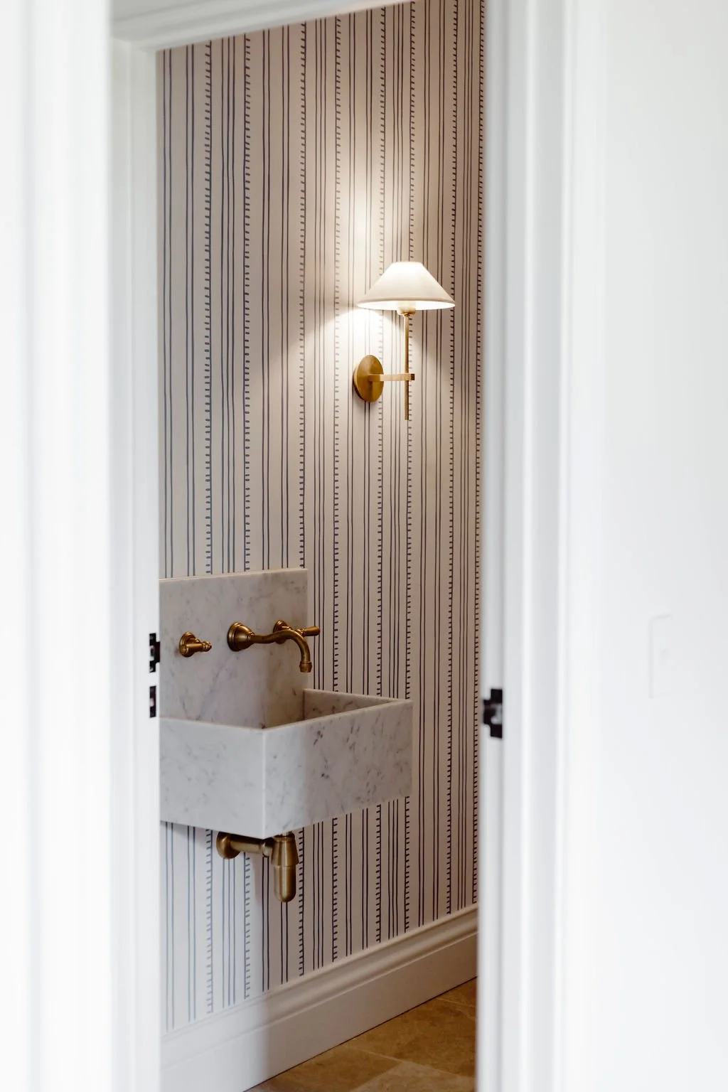 Interior view of a bathroom through a doorway, featuring a wall-mounted brass lamp, a patterned wallpaper with vertical stripes, a small marble sink with brass taps, and a beige floor.