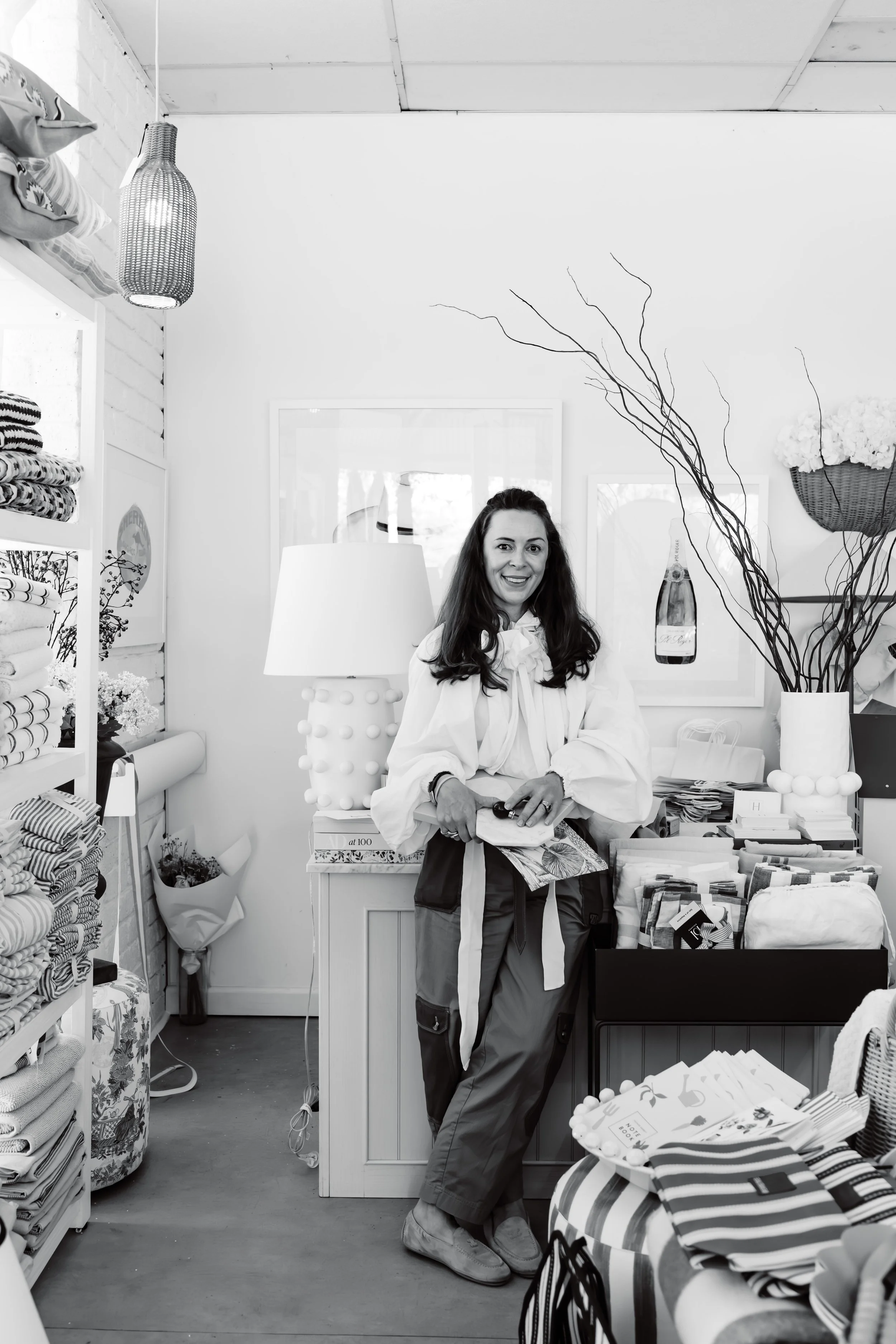 A woman smiling inside a home decor or fabric store, standing behind a counter with folded textiles and fabric swatches. Shelves with neatly organized fabrics are on the left, and decorative items such as vases, a lamp, and a large basket with flowers are visible around her.
