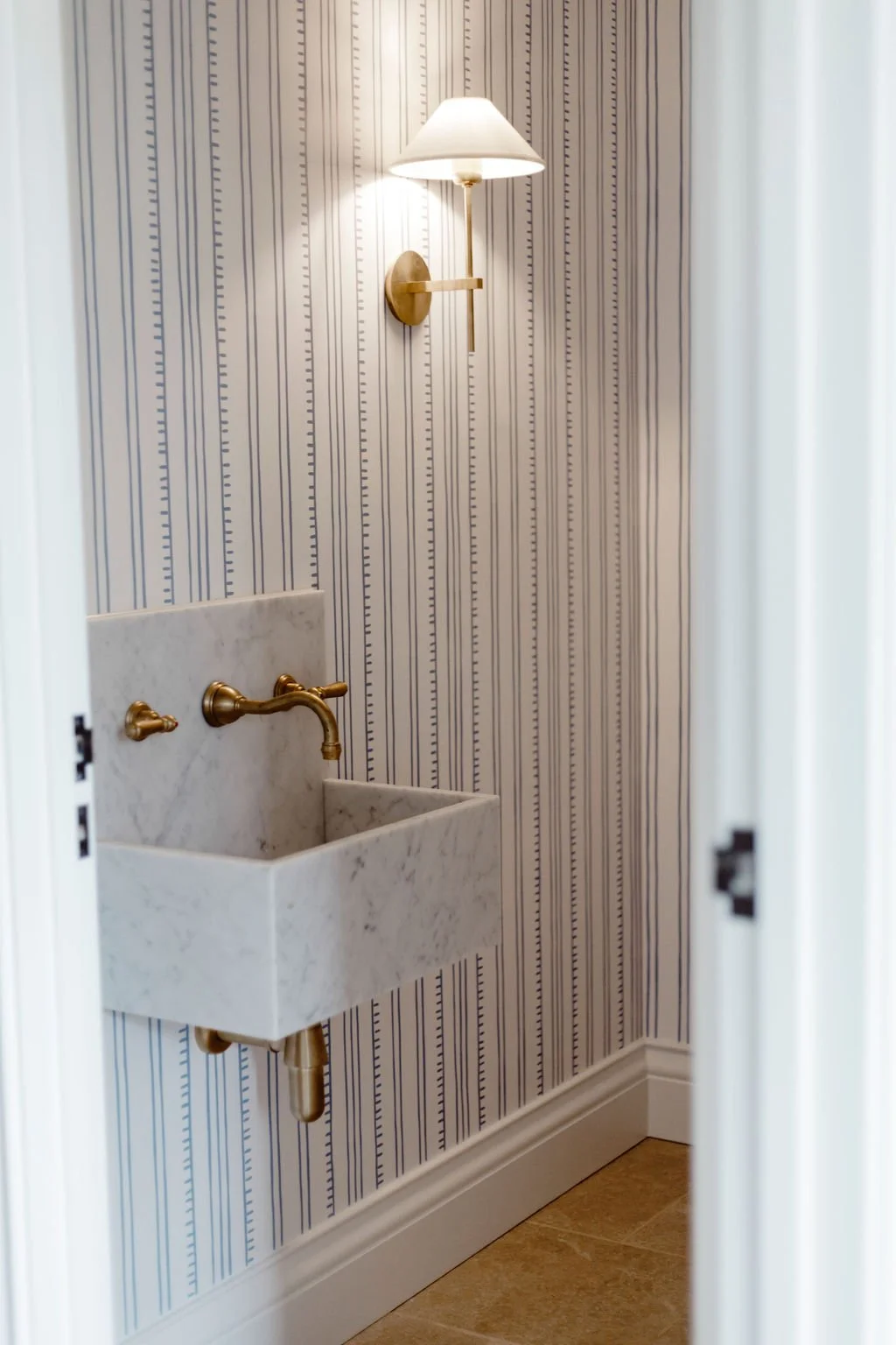 Small bathroom with striped wallpaper, wall-mounted light fixture with a shade, vintage style faucet above a small marble utility sink.
