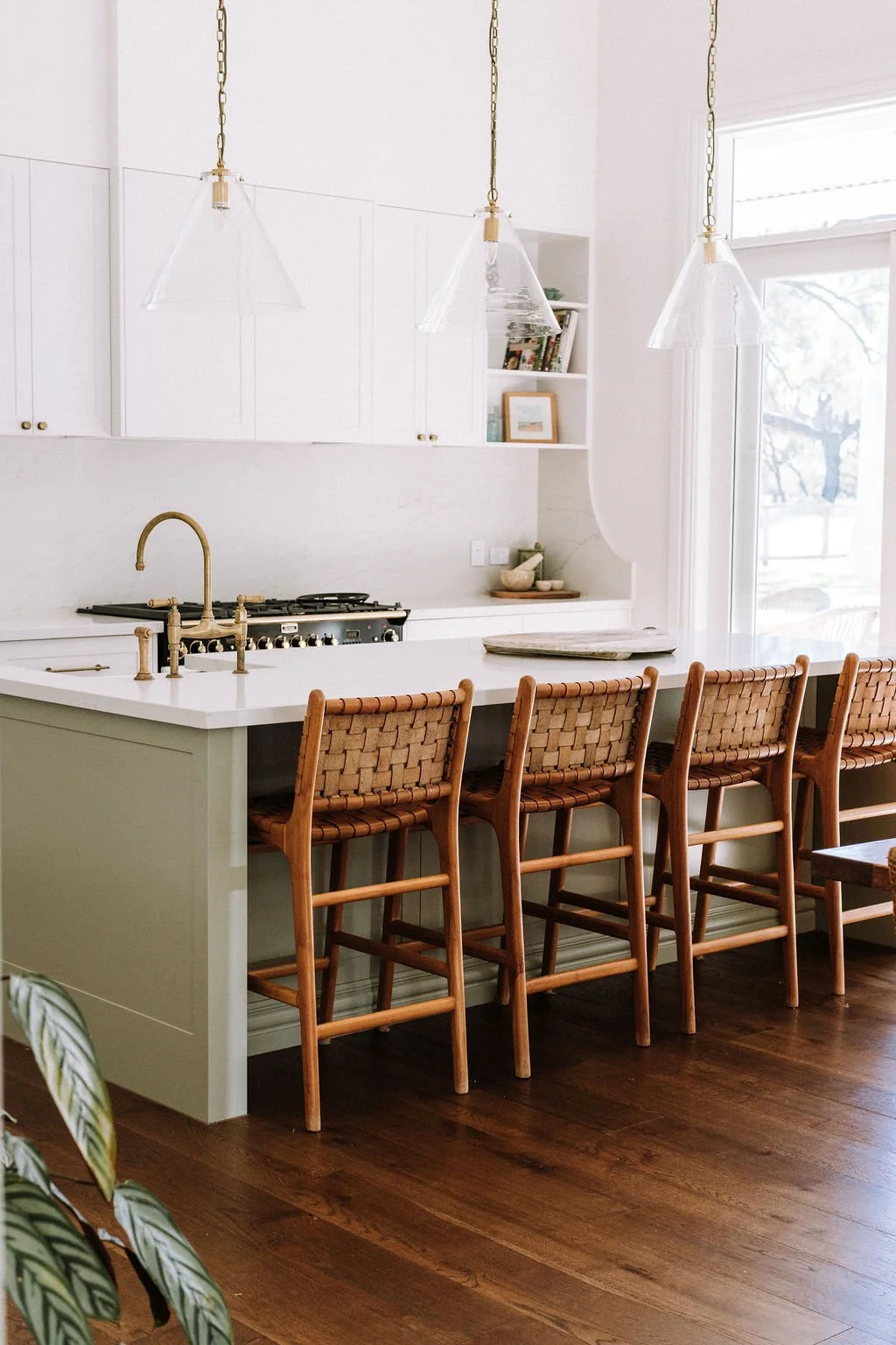 A modern kitchen with a white island featuring a brass faucet, surrounded by four woven wood chairs, and a black stovetop against white cabinets with gold knobs. There are pendant lights hanging above and a large window on the right.