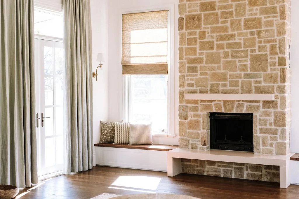 Bright living room with large windows, beige curtains, a stone fireplace, and a window seat with decorative pillows.