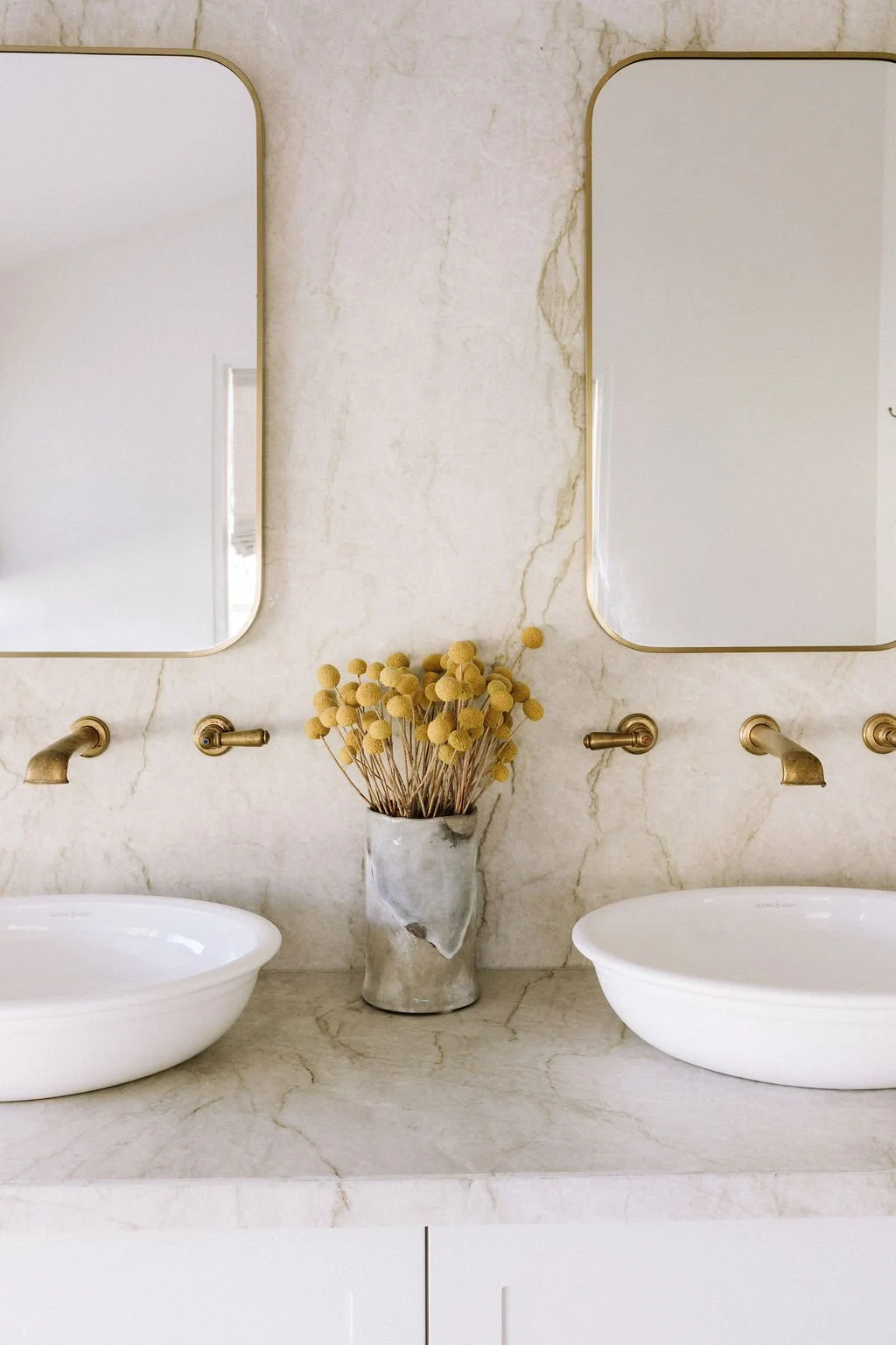 A bathroom vanity with a marble countertop, two white vessel sinks, and two mirrors above. A plant with yellow spherical flowers in a marble vase is centered between the sinks.