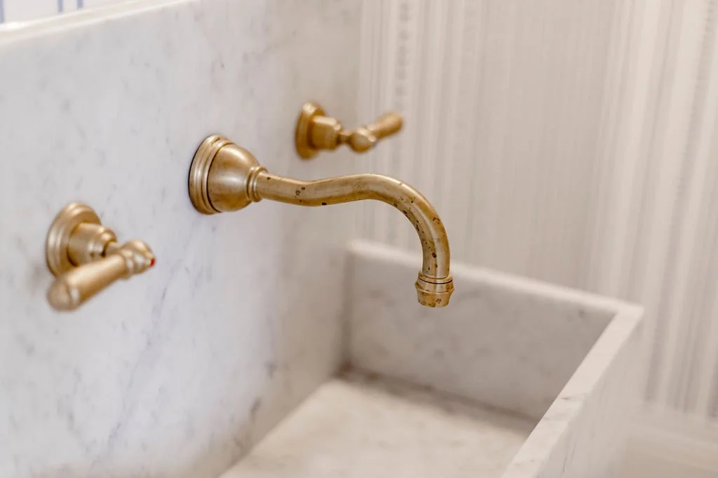 A close-up of a vintage gold-colored wall-mounted sink faucet with two handles, one for hot and one for cold water, against a marble wall background.