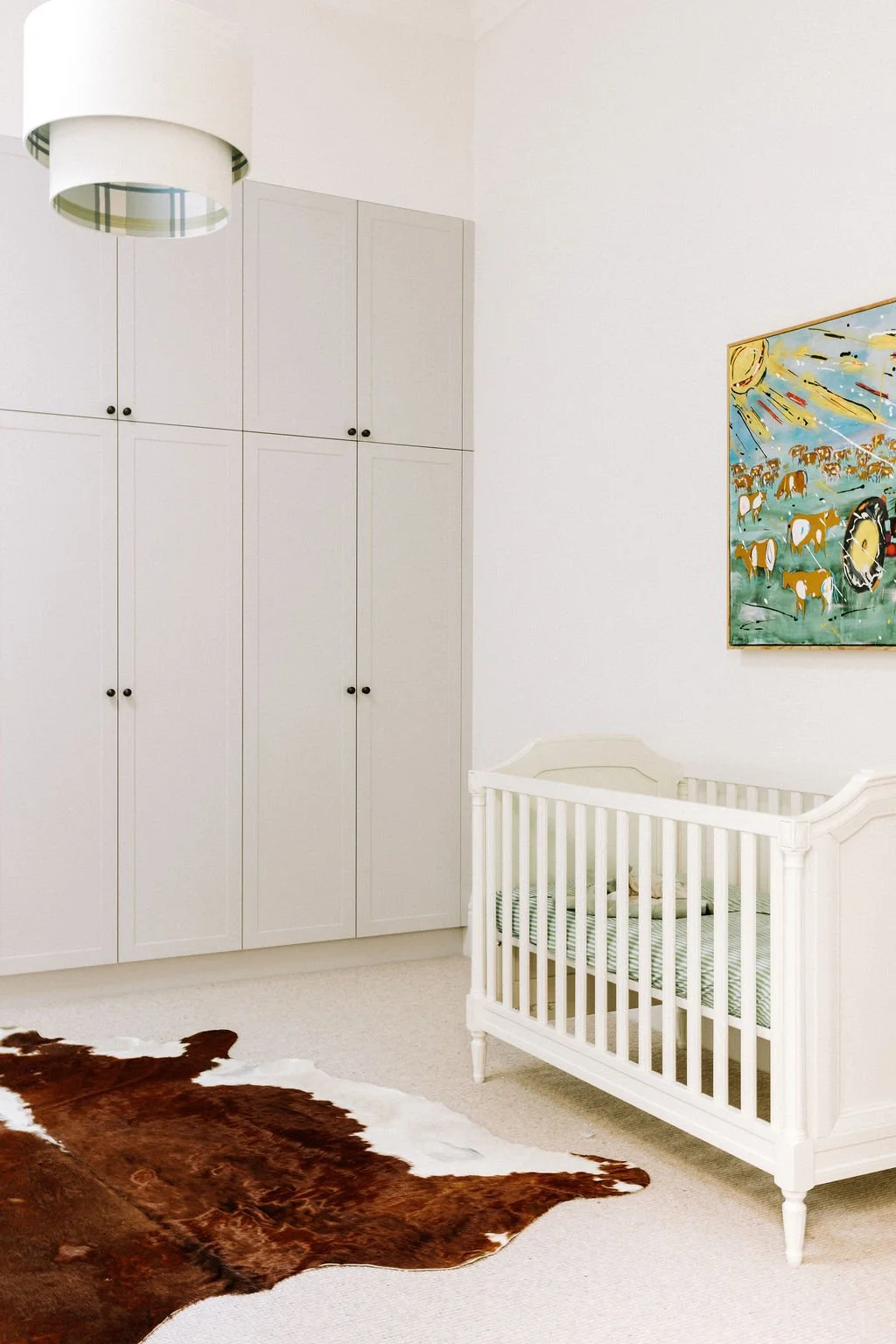 A nursery with white cabinets, a white crib, a colorful abstract painting on the wall, and a cowhide rug on a beige carpet.