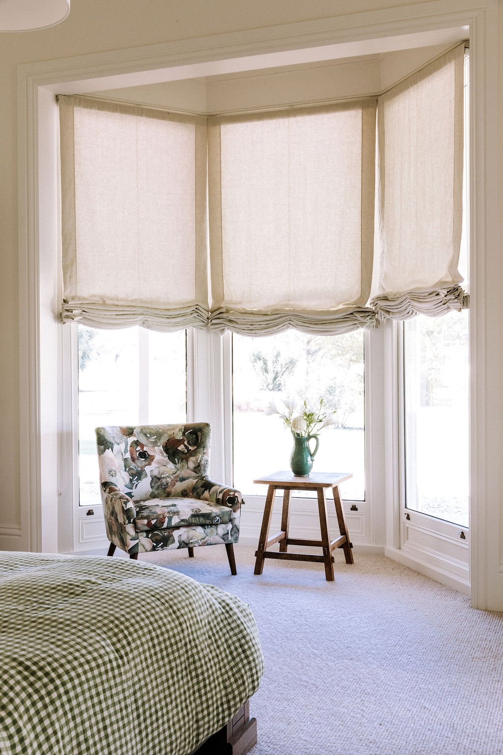 A cozy corner in a room with a large bay window covered by beige Roman shades. There is a floral armchair, a small wooden side table with a green vase and flowers, and a bed with a green gingham bedspread.