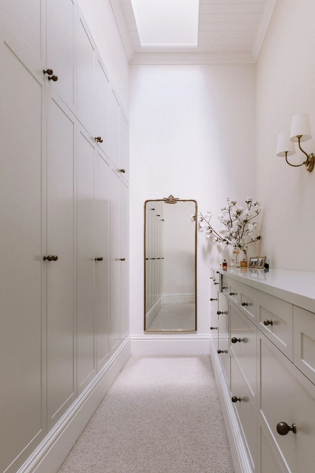 A bright, narrow dressing room with built-in white cabinets on the left, a white dresser with drawers on the right, decorated with a vase of cotton branches, framed photos, and small perfume bottles. A tall, ornate mirror leans against the wall at the end, and a small window filters in natural light.