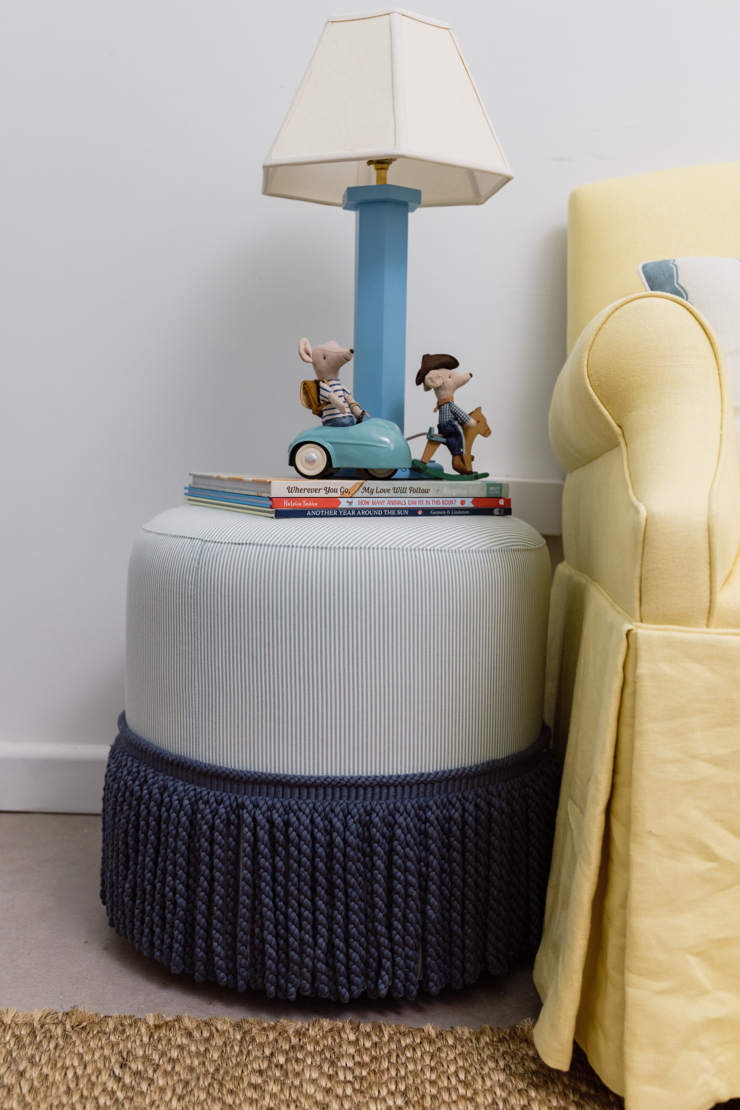 A table with a pile of books on top, a blue table lamp with a white lampshade, and two plush toys resembling mice in striped shirts. A yellow armchair is partially visible to the right.