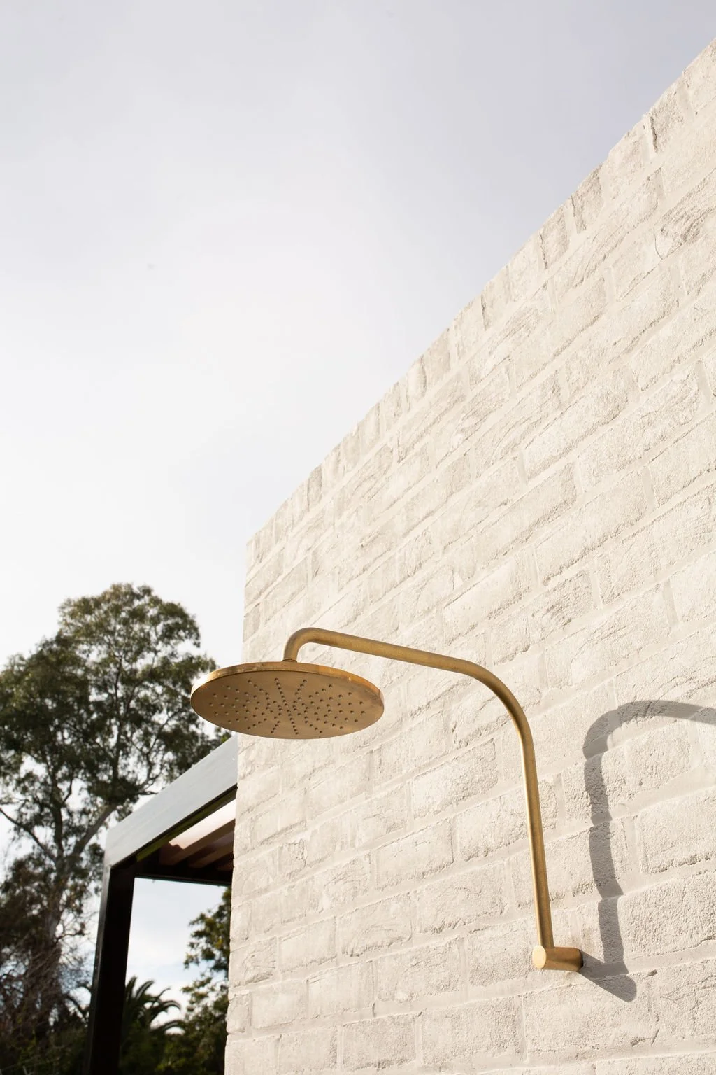 Outdoor shower with a brass showerhead mounted on a white brick wall, with a tree and cloudy sky in the background.